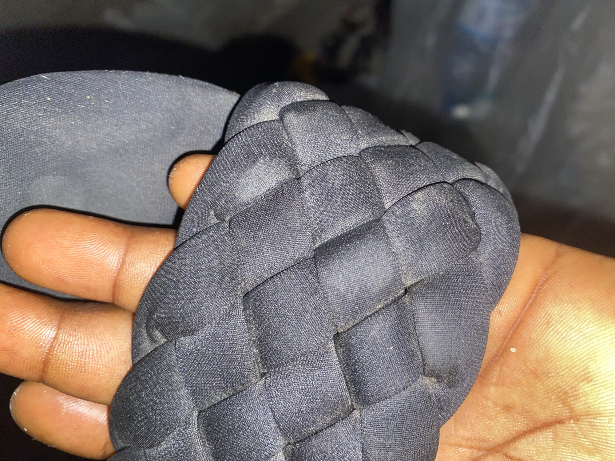 A close-up, overhead shot shows a person's hand and fingers cradling a woven black sandal in Jauro Yinu, Nigeria. The sandal's straps are wide and tightly woven, creating a textured surface. The hand's skin is a deep brown, with visible fingerprints and some light sheen, suggesting it might be slightly moist. The background is dark and out of focus, making the sandal and hand the primary subjects.