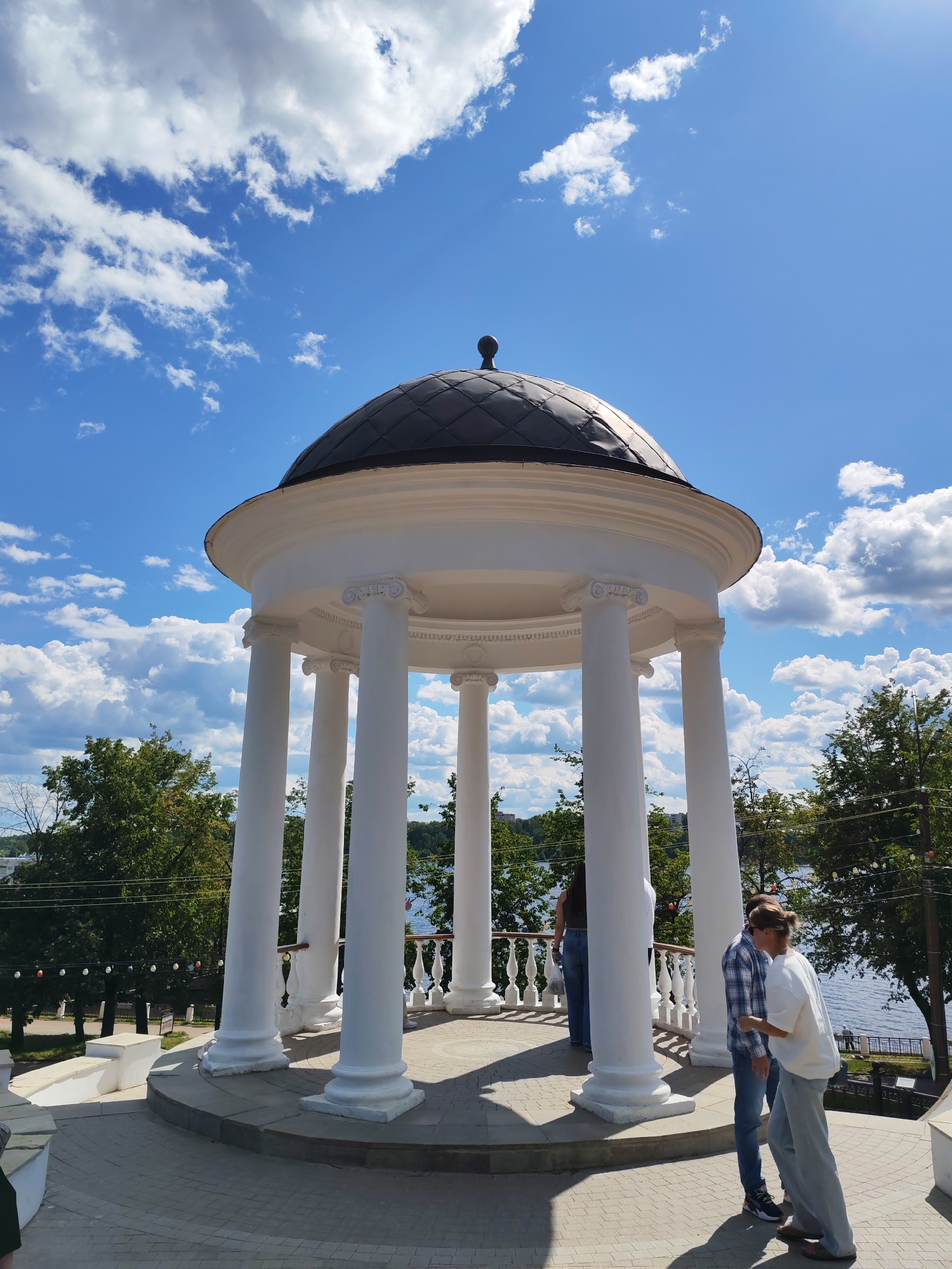 This is a photograph of the Rotunda on the embankment in Kostroma, Russia, on a sunny day with a blue sky and scattered white clouds. The rotunda is a white circular structure with a dark, domed roof, supported by several fluted columns. In the foreground, two individuals are standing near the rotunda. One person, wearing a plaid shirt and jeans, is looking down and to the right. The other person, in a white t-shirt and light-colored pants, is facing away from the camera and appears to be talking to the first individual. Behind the rotunda, a body of water, likely the Volga River, is visible, lined with trees and a walkway. A few distant figures are present on the embankment across the water. The ground in front of the rotunda is paved with brickwork.