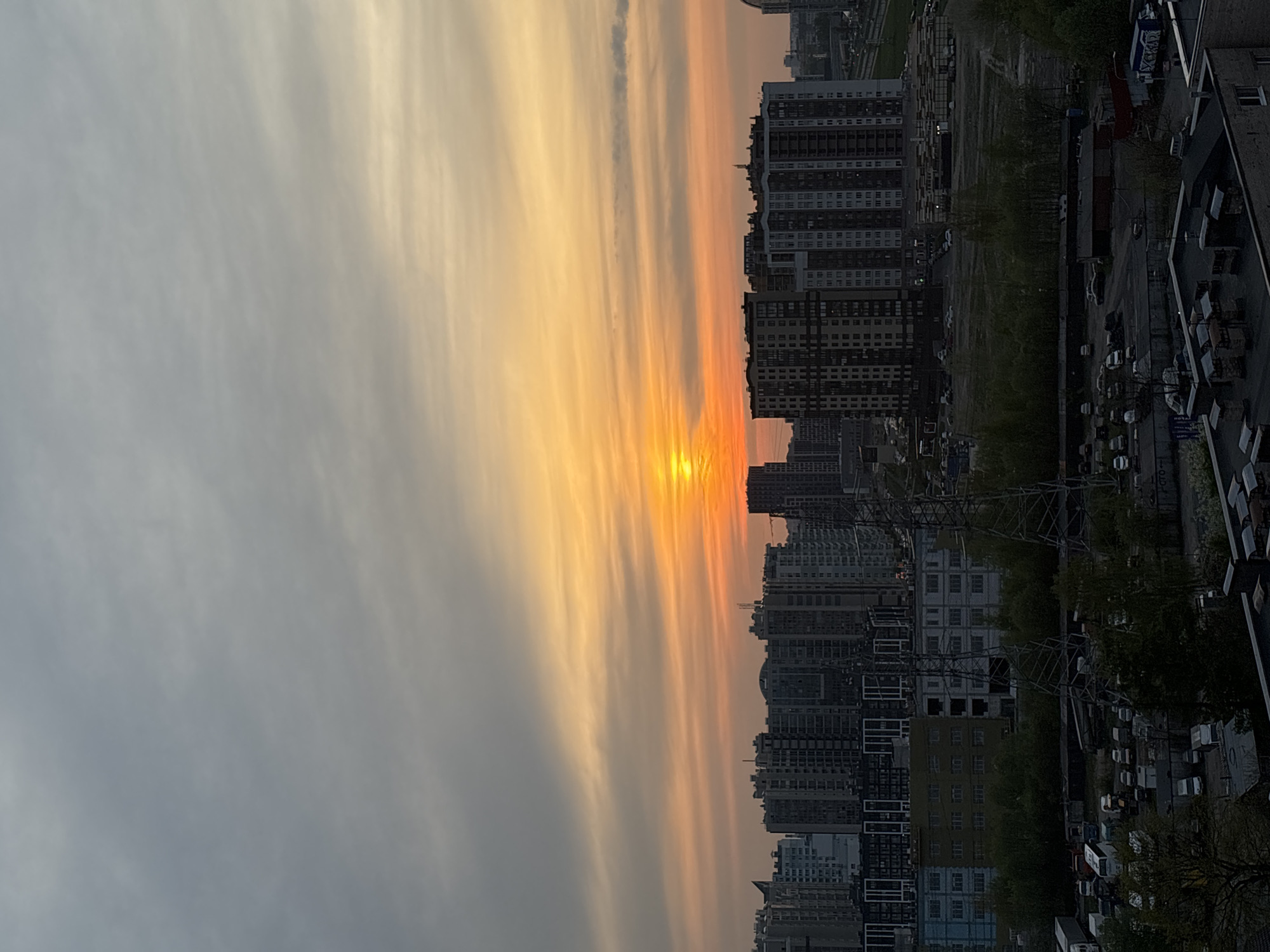 The image depicts a cityscape at sunset. The sky is a gradient of colors, transitioning from gray to orange as it nears the horizon. The sun, obscured by clouds, casts a bright orange glow. High-rise buildings line the horizon, appearing as dark silhouettes against the colorful sky. The built environment includes infrastructure and parked vehicles, suggesting a densely populated urban area.