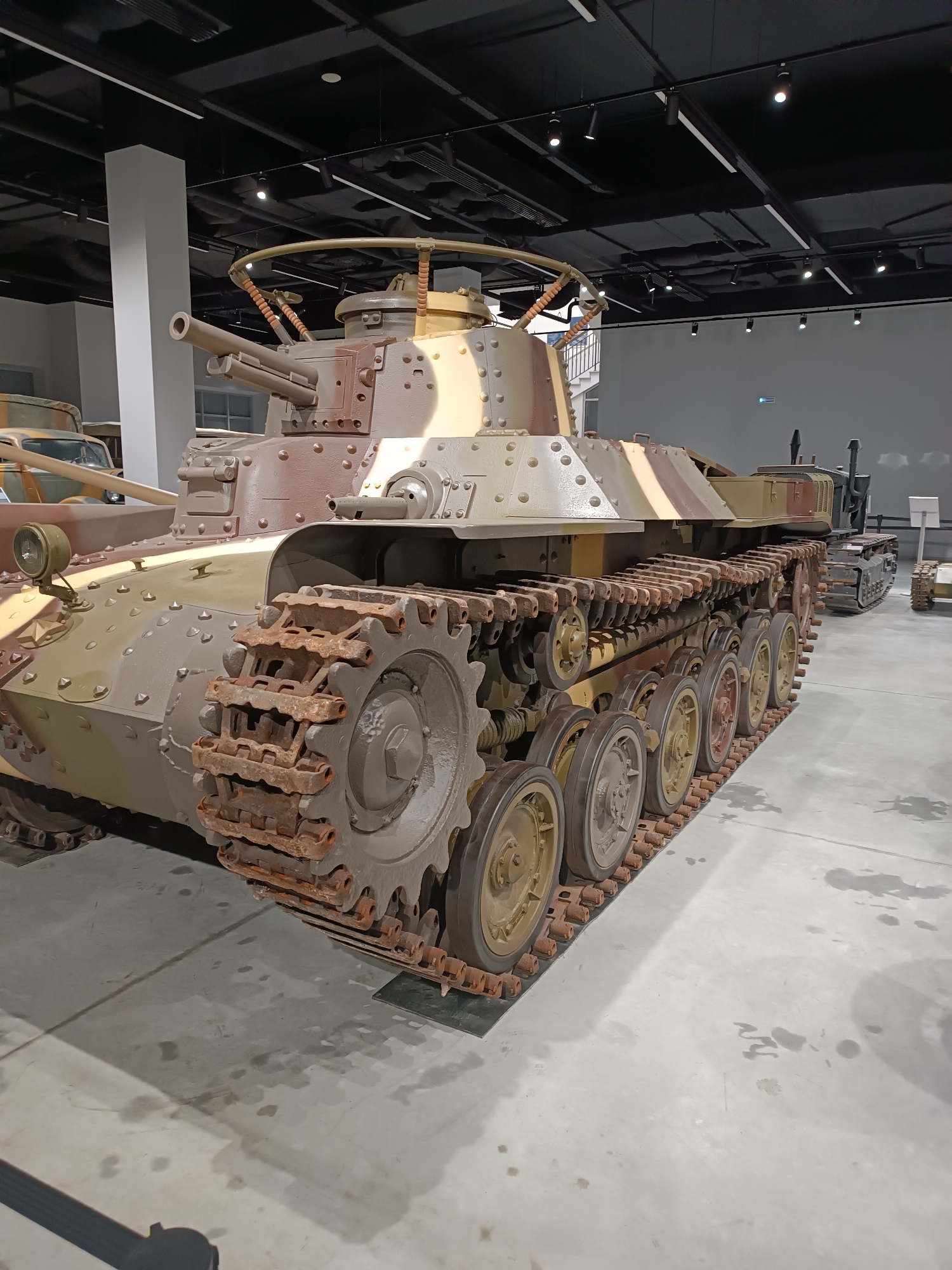 The image, taken in the city of Verkhnyaya Pyshma, Russia, depicts a museum display featuring a World War II-era Japanese Type 97 Chi-Ha medium tank. The tank is positioned on a concrete floor within a dimly lit exhibition hall. The tank, painted in a camouflage pattern of brown and tan, exhibits signs of wear and rust, particularly on its tracks and road wheels. The foreground is dominated by the tank's track system, showing numerous individual track links and multiple road wheels with yellow hubs. The tank's turret is visible, featuring a main gun and what appears to be a machine gun mount. In the background, other military vehicles, including what looks like a truck and another tracked vehicle, are partially visible. The ceiling of the hall features a grid of black beams and white light fixtures. The overall environment suggests an indoor museum setting focused on military history.