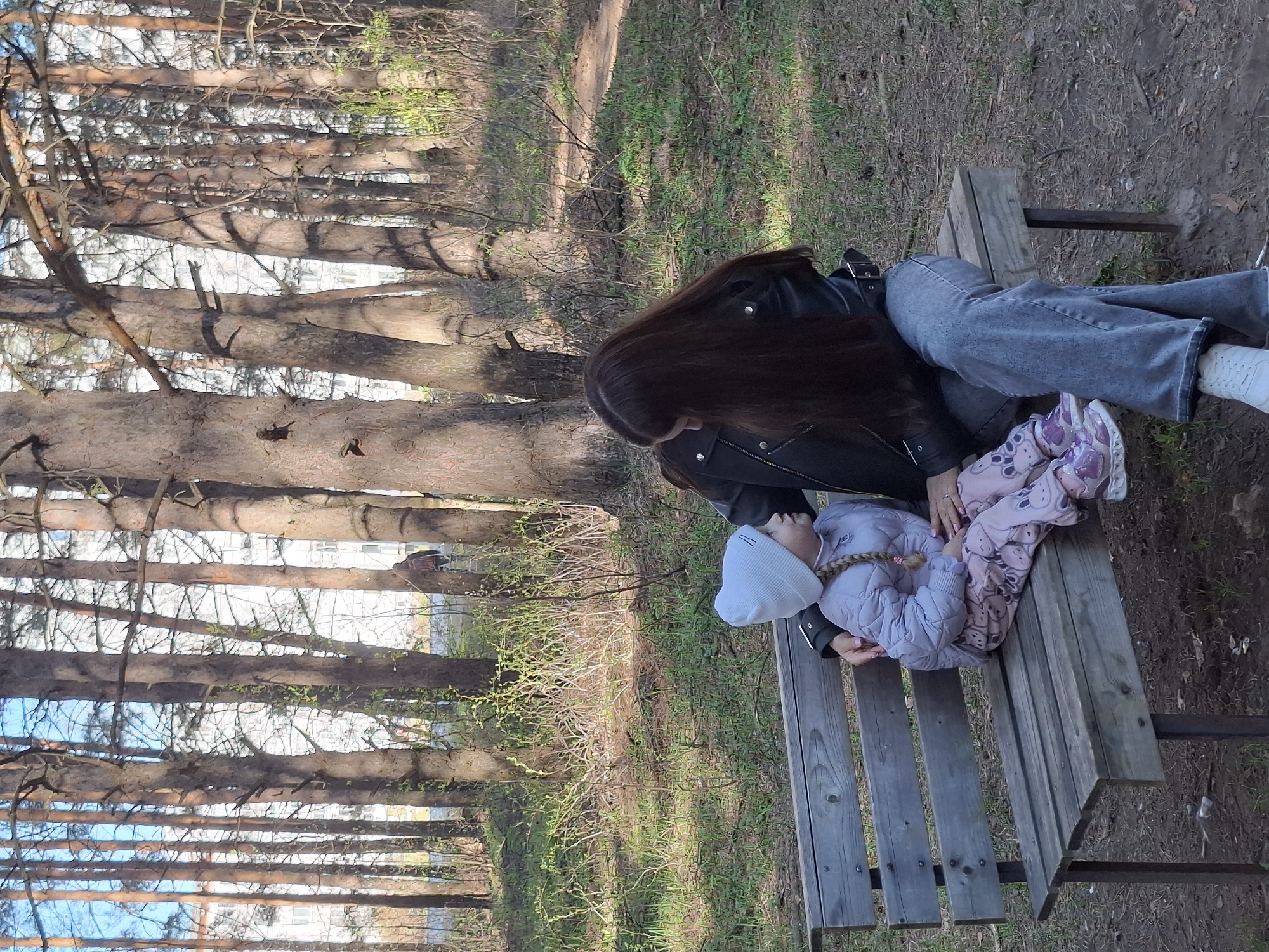 A photo shows a woman and a child on a bench in a park in Chaikovsky, Russia. The woman is seated and wearing a black leather jacket and light gray jeans. She has long, dark brown hair that falls down her back. The child is lying on the bench next to her, wearing a light purple jacket and a white knit hat. The bench is wooden and located in a grassy area surrounded by trees. Some buildings are visible through the trees.