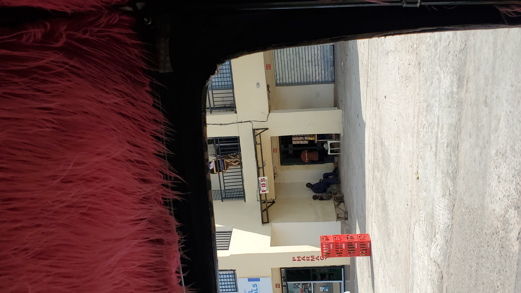 The image shows a street scene in Jalingo, Nigeria, viewed from inside a vehicle with a pink furry interior lining. In the foreground, a stack of red Coca-Cola crates is visible. Behind the crates is a cream-colored building with multiple storefronts. One storefront, labeled "PHARMACY," is on the left. To the right of the pharmacy, two men are sitting on wooden benches outside a storefront with a sign that reads "AVAILABLE POS." Another man is standing inside this doorway. Further to the right, a third storefront has a metal roll-up door. There are balconies on the second floor of the building, with people standing on them. The ground is paved concrete, with some tire marks and debris visible. The overall impression is of a commercial area on a sunny day.