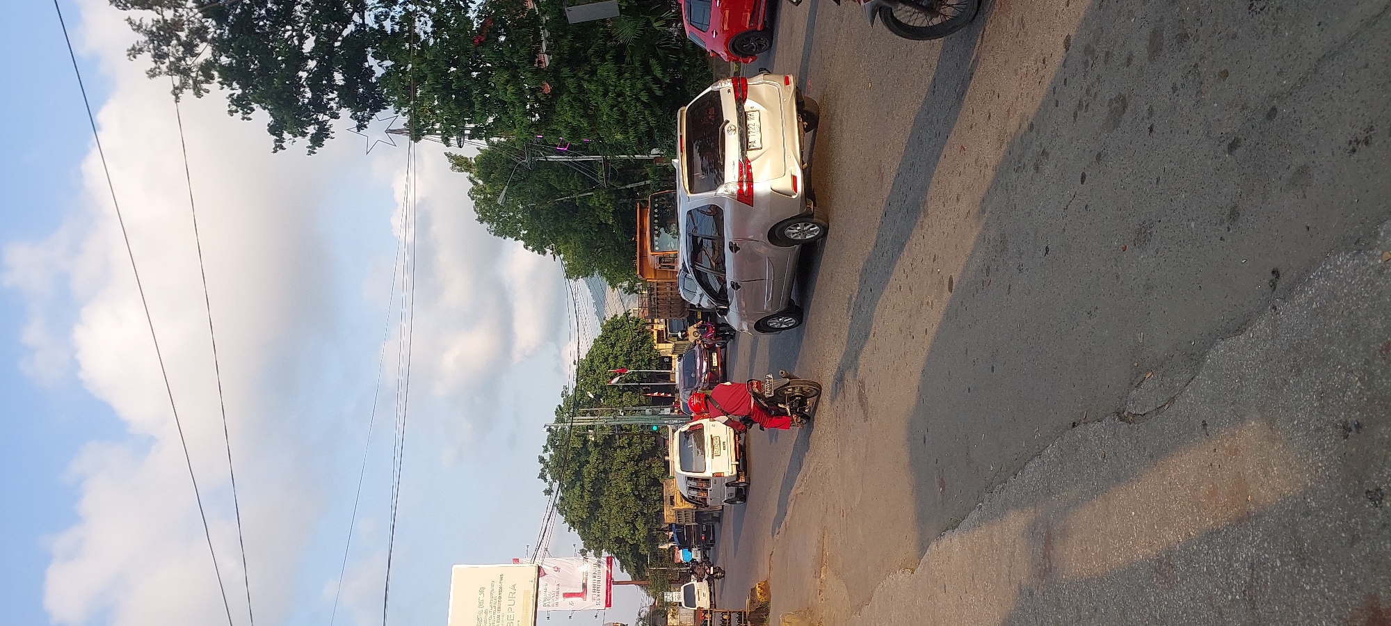 This is a street scene in Jayapura, Indonesia, likely during daylight hours given the bright sky. The road is paved and appears to be a busy thoroughfare with multiple lanes of traffic. Several vehicles are present, including cars, vans, trucks, and motorcycles.
In the foreground, a person wearing a red helmet and red clothing rides a motorcycle, heading away from the camera.
Further down the road, a silver van is prominent, with a license plate visible. To its right, a red car is parked or moving. In the background, more vehicles are visible, including trucks, suggesting ongoing traffic.
On the left side of the road, there's a billboard with text and images. Trees, some of which are quite large, line the sides of the road, and in the distance, hills can be seen. Power lines are strung overhead, supported by poles. The sky is partly cloudy with a clear blue sky visible in some areas. The lighting suggests it might be late afternoon due to the long shadows cast on the road.