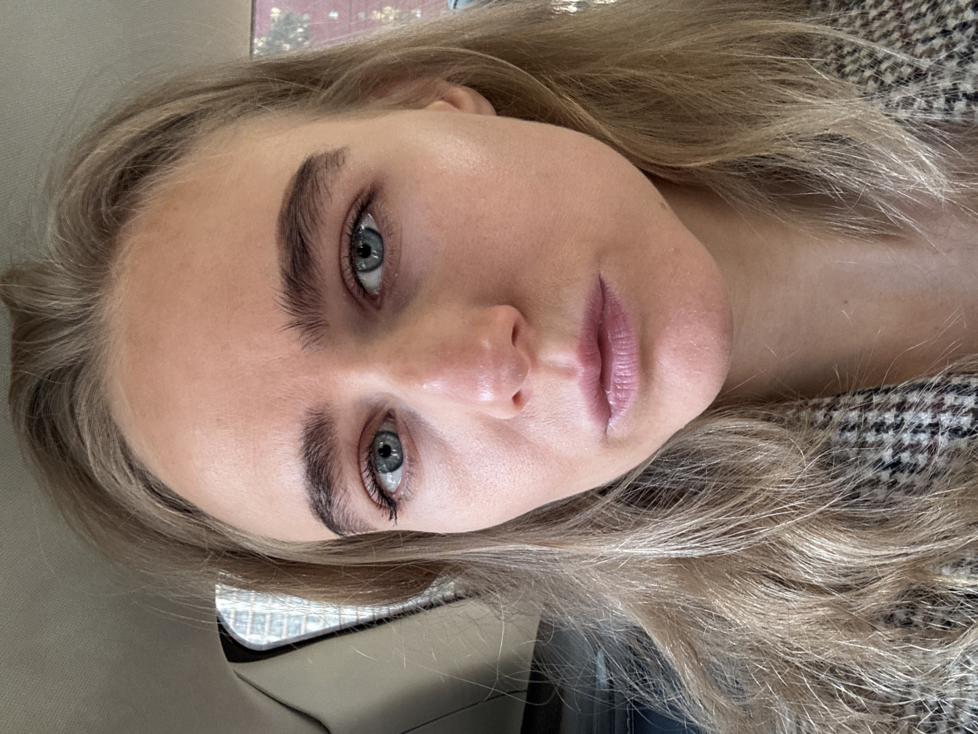 A close-up, tilted portrait of a fair-skinned woman with light blonde hair and blue eyes. She has full lips and well-defined eyebrows. Her skin appears smooth and hydrated. She is wearing a patterned blazer or jacket. The background includes a car window, a car interior, and a glimpse of buildings outside the car. The location appears to be an urban environment.