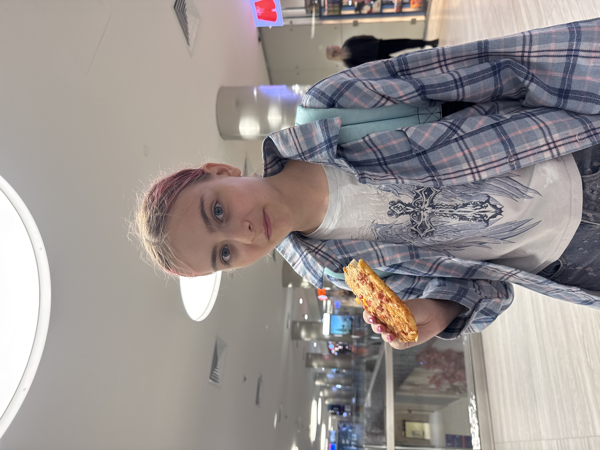 A young female with pink-streaked blonde hair, wearing a plaid shirt over a graphic t-shirt and jeans, holds a slice of pizza in her right hand and faces the camera. She has bright blue eyes and a neutral expression. She is wearing a light blue backpack. The background appears to be an indoor public space, possibly a shopping mall or airport terminal, with modern architecture, bright lighting, and visible signage in Russian. The location is Moscow, Russia.