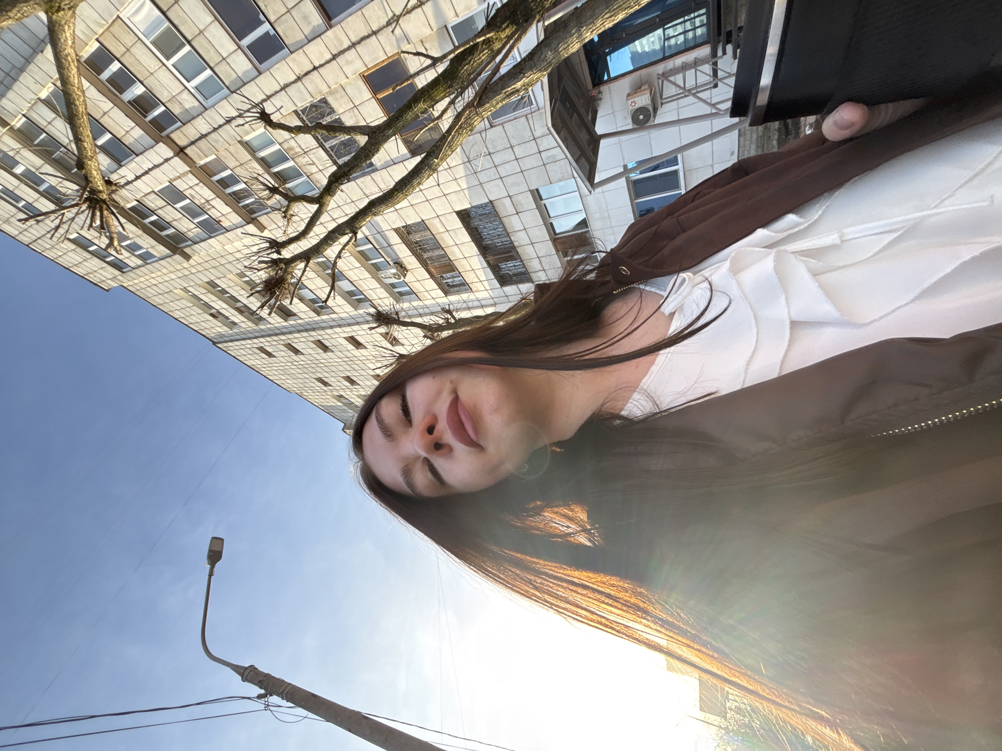 The image is taken from a low angle, looking up. A woman with long, dark hair is positioned in the center, facing the camera with her eyes closed. She is wearing a white top and a brown jacket. Above and behind her is a large building with a grid pattern of windows, partially obscured by bare tree branches. To the left, a blue sky is visible with a street lamp and power lines in the foreground. The sun is positioned low, creating a lens flare effect near the bottom of the frame. Based on the architecture, urban setting, and provided location information, this scene is likely in Perm, Russia.