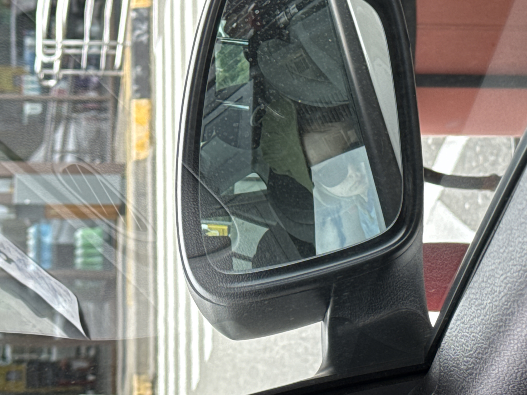 The image shows the side mirror of a vehicle, reflecting the interior of the car and elements of the outside environment. The environment visible through the windshield appears to be a commercial area, possibly a shop or store, with shelves stocked with items. The location is unknown.