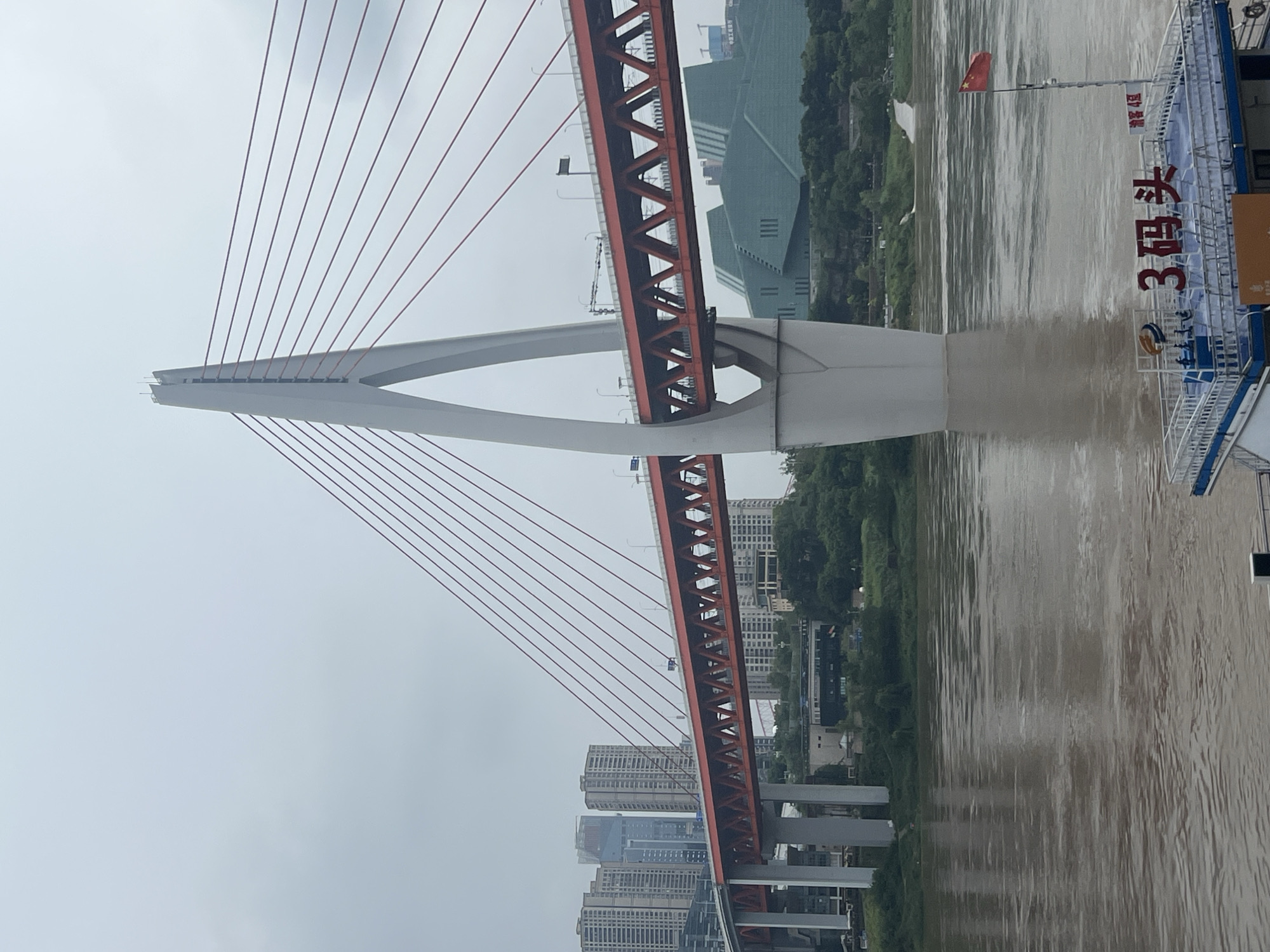 An extreme long shot captures a cloudy day over the Yangtze River in Chongqing, China. The Hongyadong Bridge, a distinctive cable-stayed bridge, dominates the frame. Its white, A-shaped tower rises from the water, supporting a vibrant red bridge deck. Numerous red cables extend from the tower, creating a striking visual pattern against the overcast sky. In the background, modern high-rise buildings line the cityscape, hinting at the urban environment of Yuzhong District. The murky brown water of the river reflects the muted light. A small boat with a Chinese flag is docked in the foreground, its deck bearing Chinese characters that translate to "3 Terminal." The overall impression is one of urban development and the power of engineering against a natural backdrop.