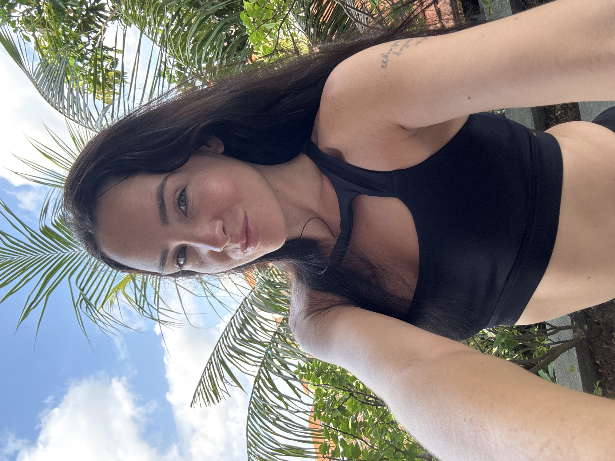 A woman with long, dark hair and fair skin takes a selfie outdoors. She is wearing a black sports bra and is looking at the camera with a slight smile. Behind her, palm fronds and lush greenery are visible against a blue sky with white clouds. The image appears to have been taken in a tropical setting, potentially in a garden or on a balcony, consistent with a location in North Kuta, Indonesia. The overall lighting suggests it is daytime.