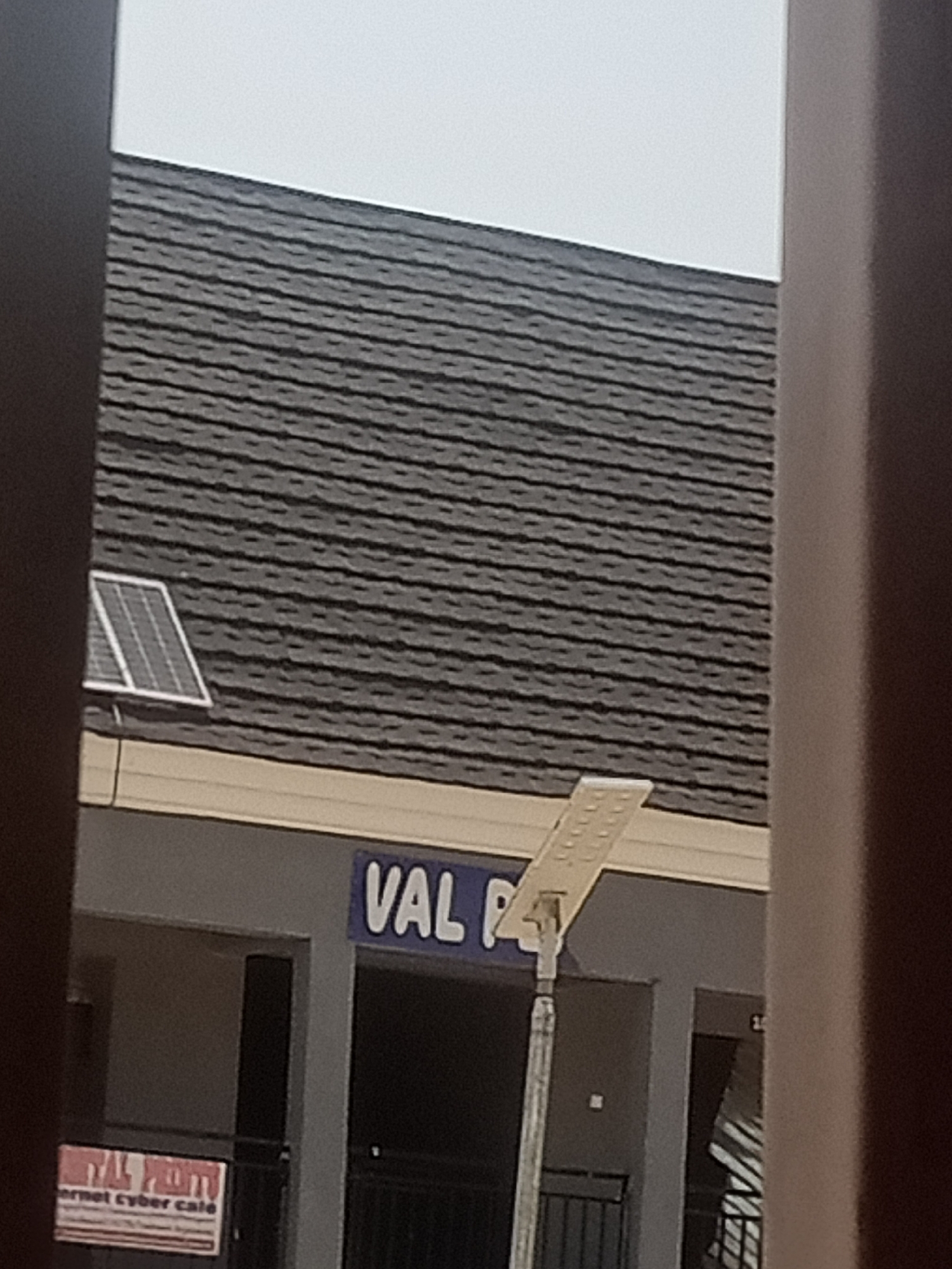 The image shows the facade of a building with a dark grey, shingled roof. A small solar panel is mounted on the roof. Below the roof, a beige trim runs along the building. Further down, a grey wall is visible, with signage indicating "VAL P" in large, blue letters. To the left, a red and white sign reads "VAL PRO" and "ernet cyber cafe". A modern street light with a metal pole and a white panel is positioned in front of the building. The scene appears to be outdoors, possibly in a commercial area, given the signage. The lighting suggests it could be daytime, though the sky is overcast, indicating potentially cloudy weather. The image is framed by dark vertical elements on the left and right, suggesting it was taken through a window or doorway. There are no people visible in the frame.