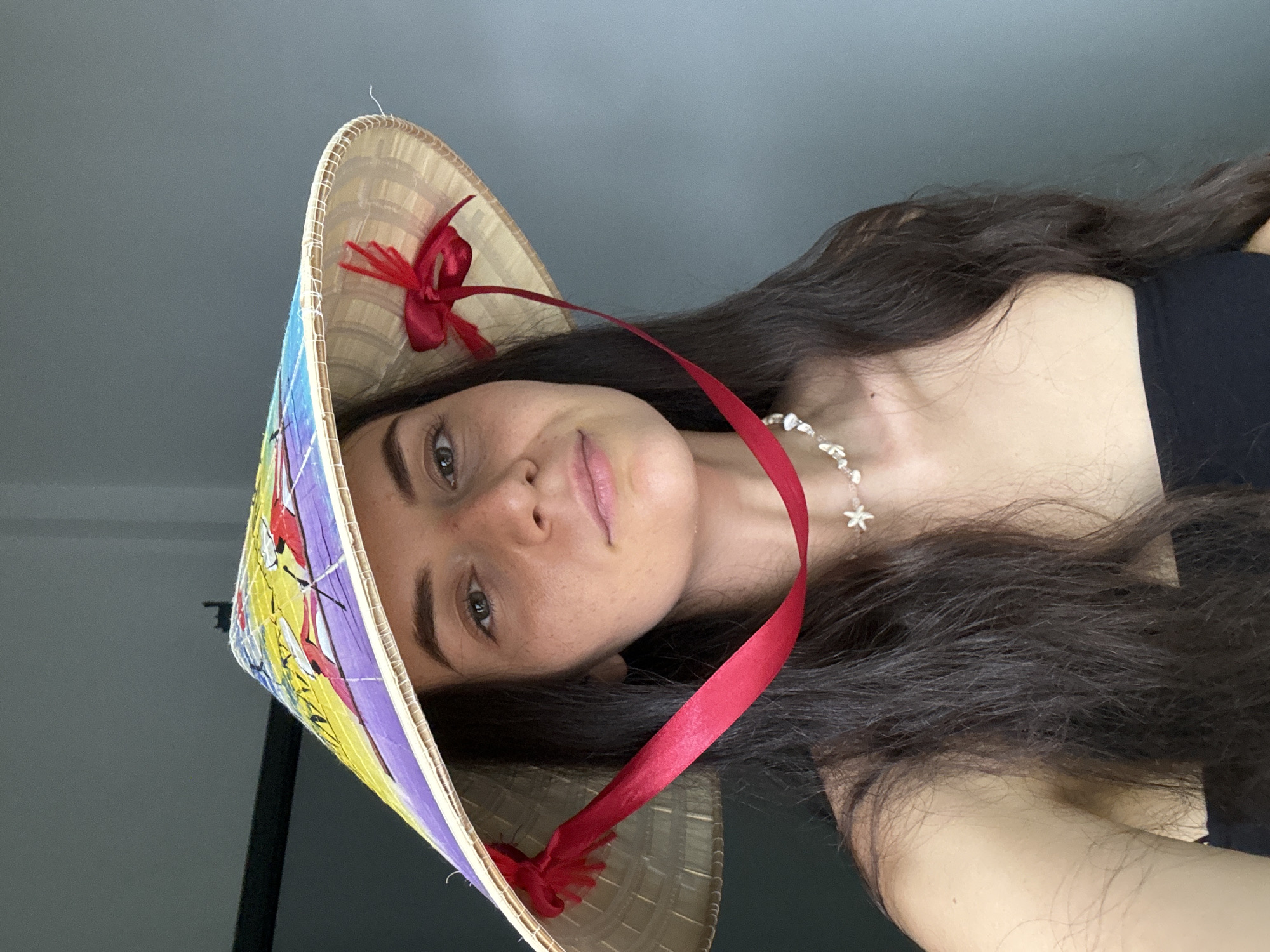 The image shows a woman wearing a Vietnamese conical hat, also known as a "nón lá." The hat is made of woven straw with a colorful design on the brim. It has a red ribbon attached to the inside. The woman has long dark hair and is wearing a necklace. The background appears to be an indoor space with gray walls. Since the city location of Mũi Né, Vietnam, was provided, and the woman is wearing a traditional Vietnamese hat, it can be inferred that the image was taken in Mũi Né.