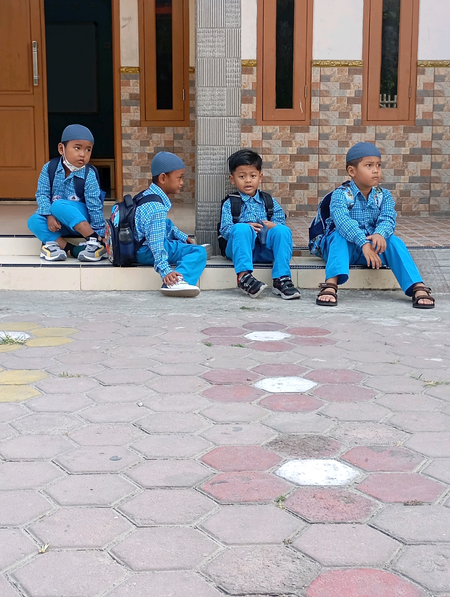 The image shows four young boys sitting on steps outside a building. They are all wearing blue checkered shirts, blue pants, and blue caps. Three of the boys have backpacks. The building has a tiled facade and wooden-framed windows. The ground in front of the steps is paved with hexagonal tiles in various shades of gray, pink, and yellow. One boy on the far left is wearing a white mask. The setting appears to be a school or religious institution in Babelan, Indonesia.