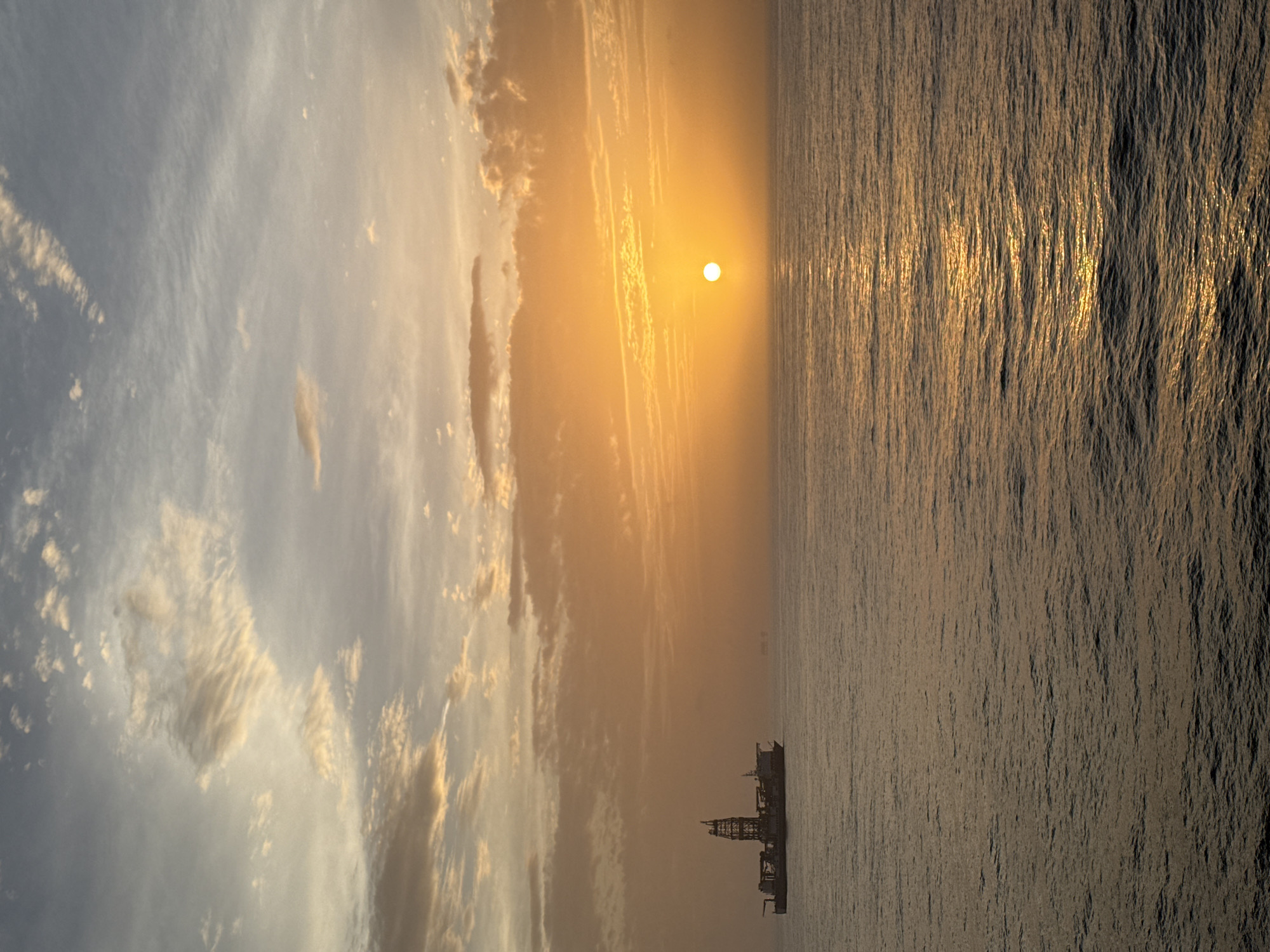 The image depicts a seascape at sunset. The sun, a bright circular orb, is positioned centrally in the frame, casting a golden light across the water. The sky is a mix of blue and golden hues, with clouds scattered across the left side. A dark, silhouetted structure, possibly an oil rig or ship, is visible in the lower center of the image. The water appears choppy, with reflections of the sun creating a shimmering effect. The location is a body of water, likely an ocean, with the structure indicating a potential offshore industrial activity. The city and specific location are not discernible from the image.