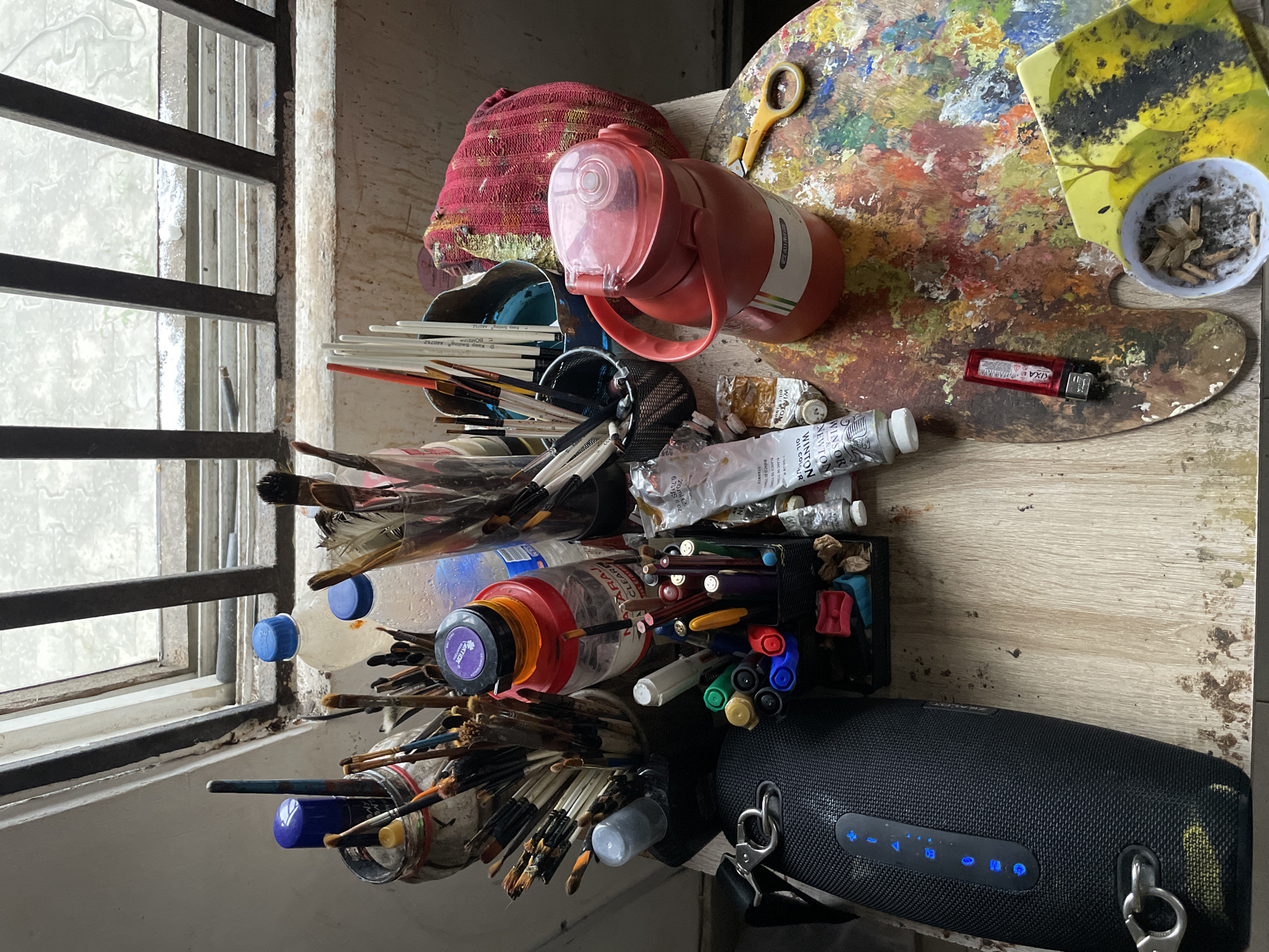 This is a high-angle, medium shot of an artist's cluttered workspace, likely located on a windowsill in Ado, Nigeria, given the presence of a bottle with "Naija Clear" branding. The scene is illuminated by natural light filtering through a window with metal bars.
Dominating the foreground is a well-used wooden artist's palette covered in a vibrant array of dried oil paint colors. Resting on the palette is a small, yellow-handled pair of scissors and a red plastic lighter. To the right of the palette is a white ashtray containing cigarette butts and ash.
To the left of the palette, a large, black portable speaker with illuminated blue buttons is prominent. A metal clip is attached to its side.
Further back, various art supplies are organized in containers. These include numerous paintbrushes of different sizes, some in a clear jar, others in a black mesh holder. Several tubes of Winsor & Newton oil color paint are visible, along with a red thermos with a clear lid and a red handle. A red and white striped cloth is draped behind the thermos. A bottle of clear liquid with a blue cap sits near some brushes.
A black pen holder contains an assortment of pens and markers in various colors, including green, yellow, red, blue, and purple. A tube of white paint with a black cap and a label reading "GRIMMIE" is also present.
The overall impression is one of an active, though somewhat messy, artistic environment, with evidence of recent work and use. The clutter suggests a creative process in motion.