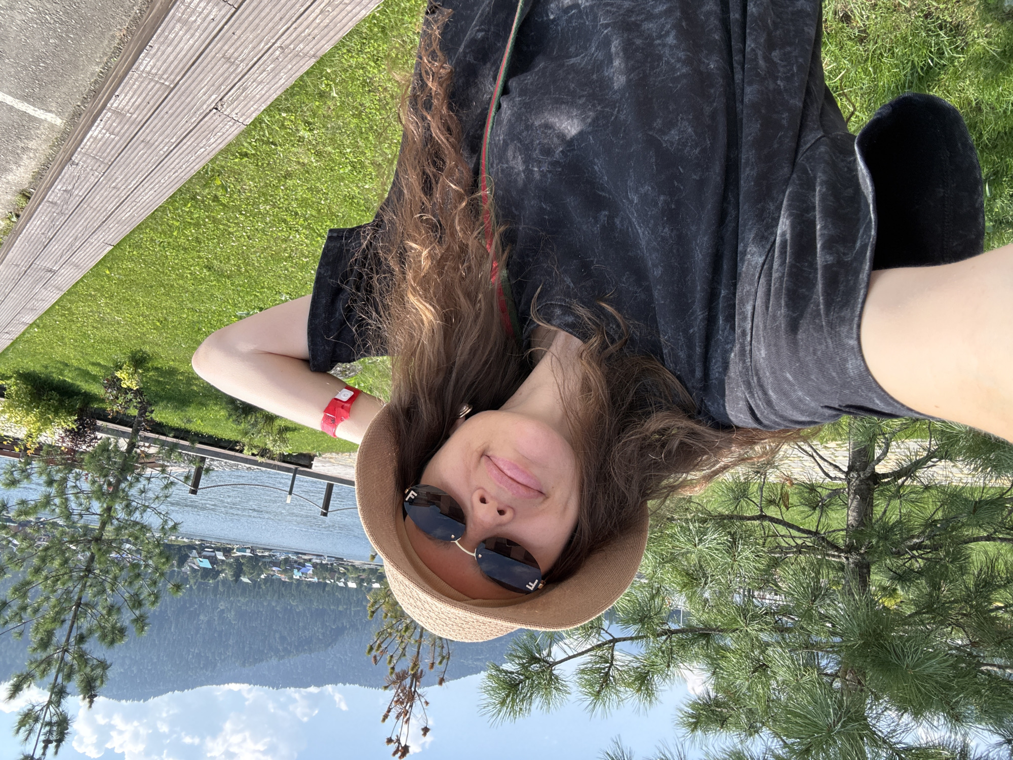 A woman is taking a selfie outdoors in Artybash, Russia. She is wearing a black t-shirt, a straw hat, and sunglasses. She has long brown hair and is smiling. In the background, there is a lake, mountains, and trees. The sun is shining and the sky is blue. She is also wearing a red wristband.