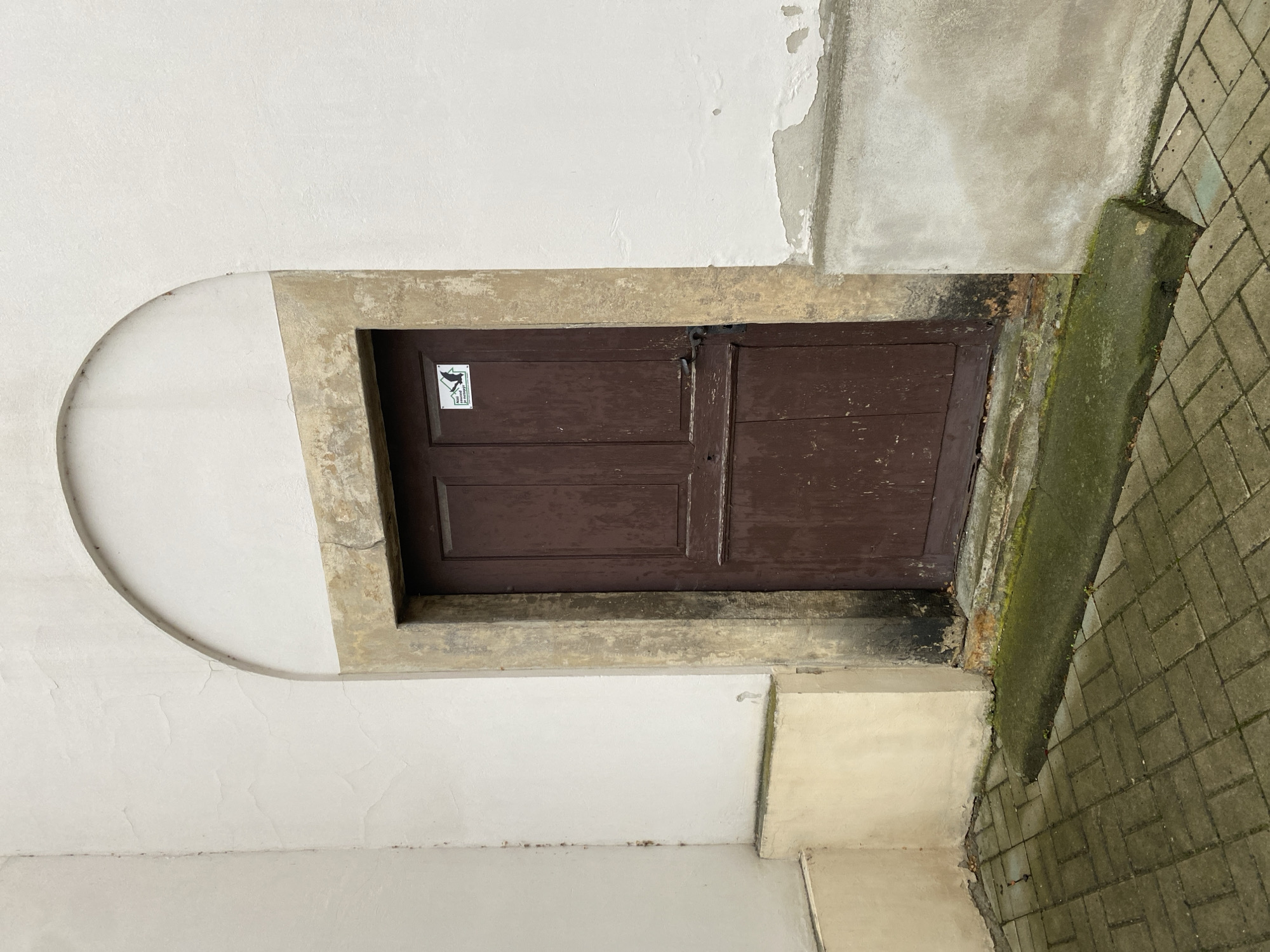 An arched opening frames a weathered, dark brown wooden door set into a cream-colored stucco wall. The door has two panels and a black metal handle. A small white sticker with a black silhouette of a bird in flight and text in an unknown language is affixed to the upper portion of the door. The door is surrounded by a stone archway that is stained and weathered. To the left of the door, the wall transitions to a lighter, creamy yellow color at the base. The ground in front of the door is paved with rectangular grey bricks laid in a herringbone pattern, with a raised concrete threshold covered in moss. The overall scene appears to be an exterior entrance to a building, possibly an older structure, located in Bílina, Czechia. The lighting suggests an overcast day.