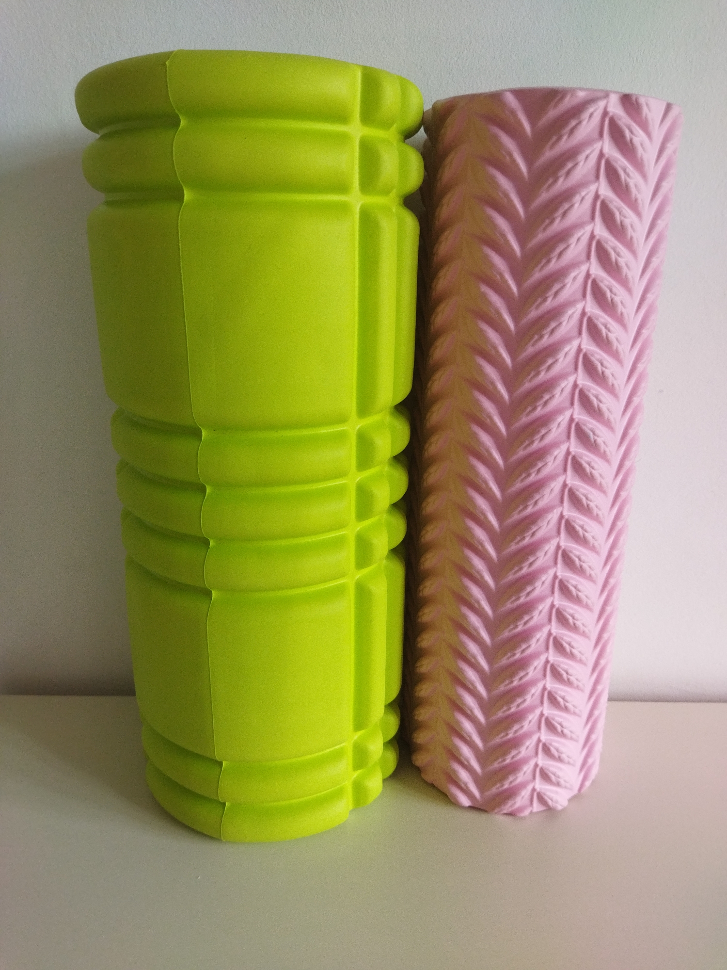 Two foam rollers are displayed standing upright on a light-colored surface against a white wall. The foam roller on the left is a bright lime green and has a segmented, textured surface with rounded protrusions. The foam roller on the right is light pink and features a raised, leaf-like pattern arranged in a chevron design. The rollers are positioned close together, with the green roller slightly in front of the pink one.