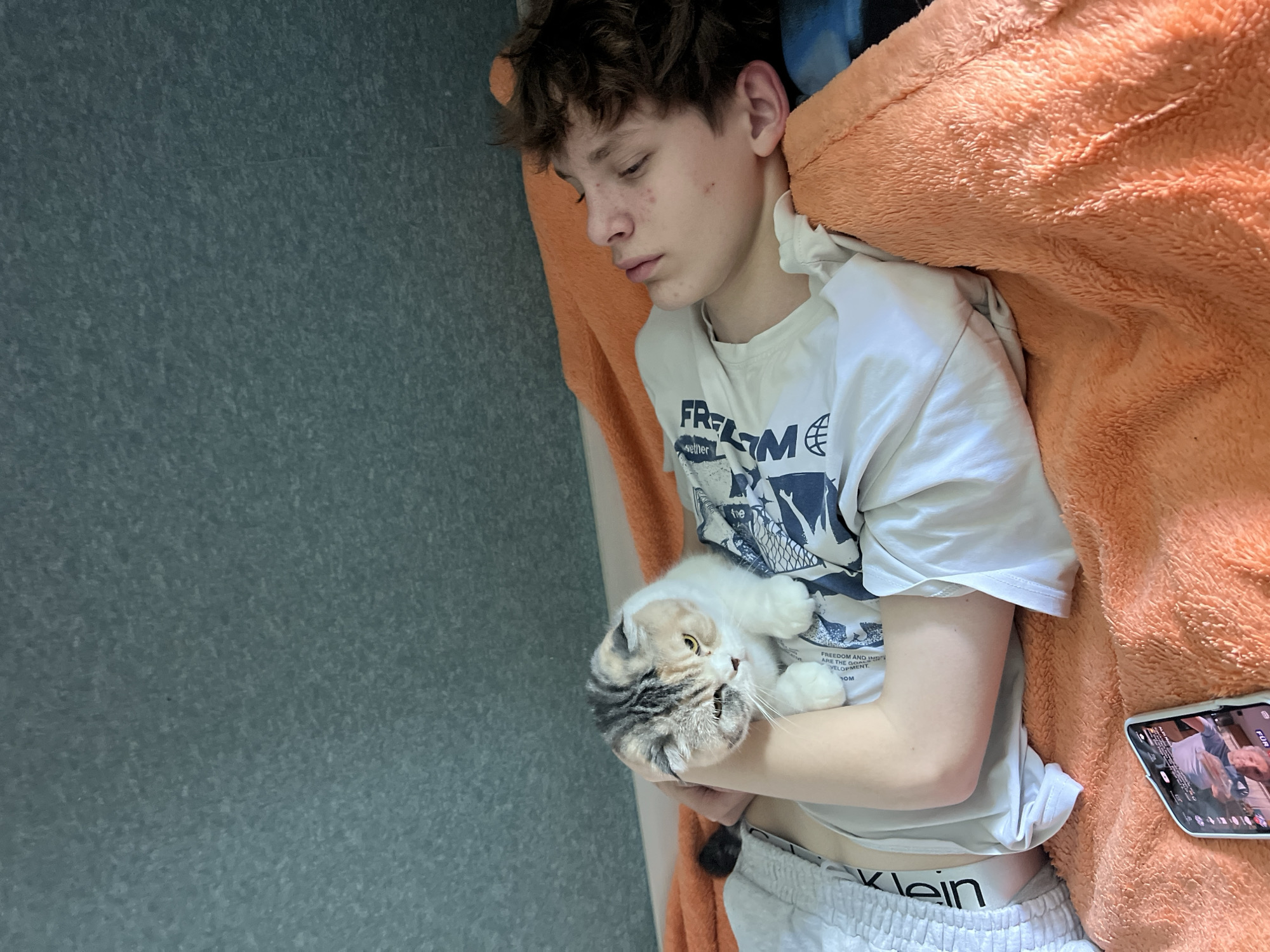 A pale-skinned adolescent male with curly brown hair lies on a bed covered with an orange blanket. He is wearing a white t-shirt and gray Calvin Klein shorts. He appears to be sleeping. A folded smartphone is resting on the blanket to his right. In his arms, he cradles a white and gray tabby cat. The background is a gray wall with a textured pattern. The location is in the city of Noviy, Russia.