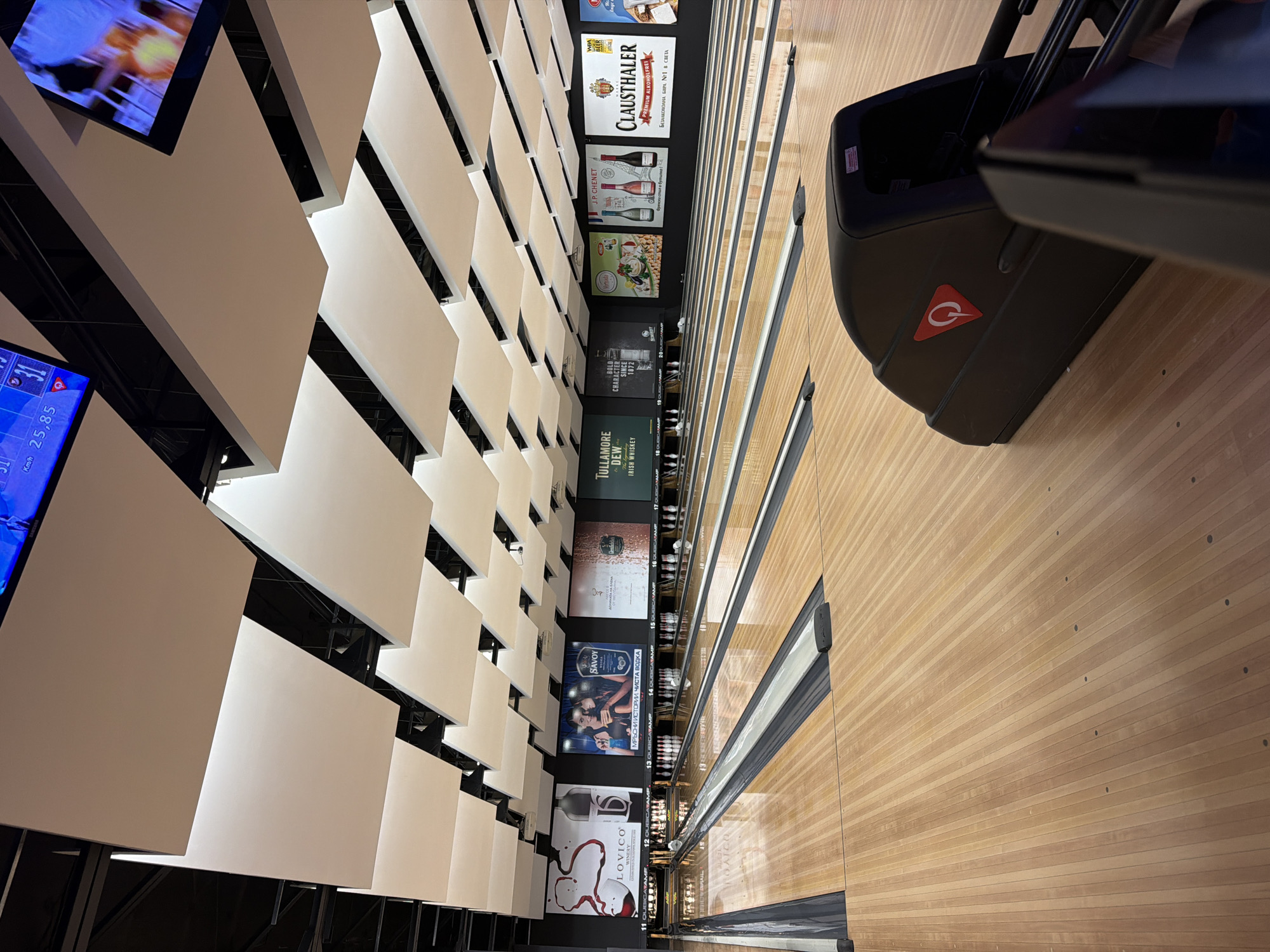 Here is a detailed description of the image:
The image shows a lane inside a bowling alley. The camera is positioned overhead, looking down the lane, angled slightly towards the wall. On the wall, there is a vertical arrangement of posters advertising various beverages, including wines, beers, and spirits, such as Lovico Winery, Savoy Vodka, Clausthaler beer, Tullamore Dew Irish Whiskey, and JP Chenet.
Next to the bowling lane, there is a ball return system in black with a prominent "Q" logo, partially obscuring the lane and possibly indicating the brand of the bowling equipment. The lane is made of wood and shows the characteristic patterns associated with bowling lanes.
The ceiling has an architectural design, consisting of white rectangular panels or slabs arranged in a stepped, staggered pattern. This design feature adds depth and visual interest to the interior space.
In the upper left corner, there are glimpses of television screens showing content, presumably related to the bowling game or providing entertainment for players.
The bowling alley may be located in the city of Sofia, Bulgaria, based on certain Cyrillic letters on one of the ads.