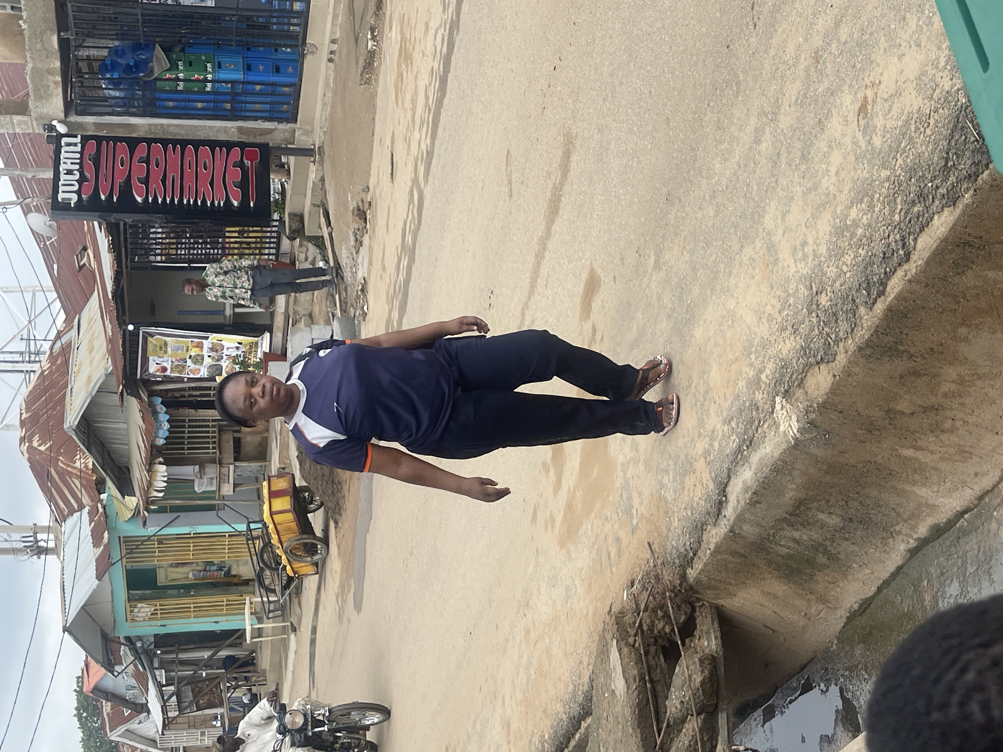 In a street scene in Abuja, Nigeria, a woman walks towards the viewer, dressed in a navy blue t-shirt with orange and white trim and navy blue athletic pants. She wears open-toed sandals. The street is paved with asphalt and has a drainage ditch to her left, which appears to be partially filled with water and debris. In the background, several small shops with corrugated metal roofs line the street. One shop has a prominent vertical sign that reads "JUCILL SUPERMARKET" in red and white lettering. A man in a camouflage shirt and grey pants stands near the supermarket entrance. A man on a motorcycle wearing a white shirt rides past in the distance. A yellow handcart loaded with yellow items is parked near one of the shops. The sky is overcast.