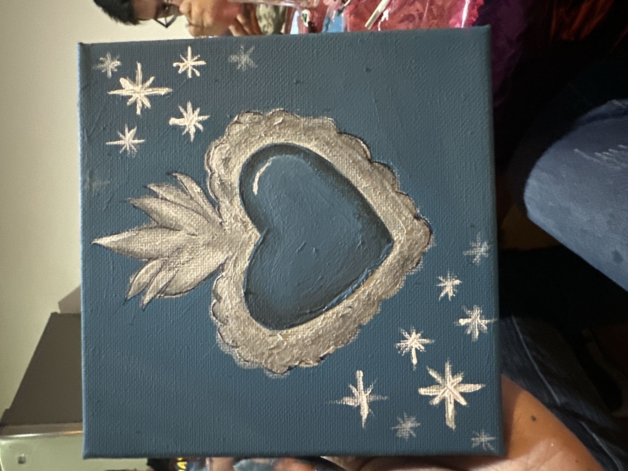 A square canvas painting features a blue background adorned with silver painted elements. A central silver heart with flames and a textured outline dominates the painting. Surrounding the heart are stylized silver star shapes of various sizes. The painting is held in a person's hand, and the edge of a table or counter is visible in the lower left corner. Other blurred figures or items are visible in the background, suggesting an indoor setting. The city and region are not identifiable from the provided image.