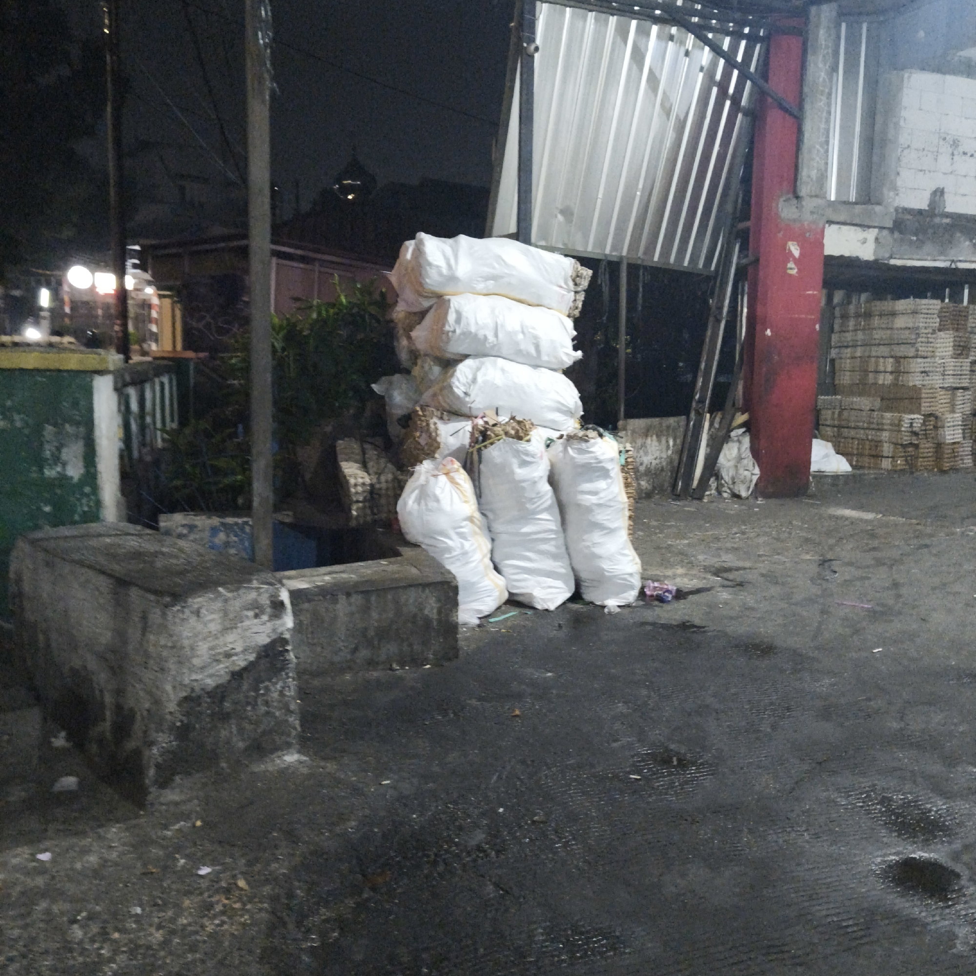 The image depicts an outdoor scene at night in a city setting, likely in South Jakarta, Indonesia, characterized by a dimly lit environment with artificial lighting. In the foreground, a concrete barrier or ledge is visible on the left, showing signs of wear. Behind this, a pile of white sacks, presumably filled with goods or refuse, is stacked and some are scattered on the ground. These sacks appear to be made of a woven material, and some have strings or ties visible.
Further back, a tall, slender, metal pole stands vertically. Behind the sacks, a corrugated metal structure, possibly part of a building or shelter, dominates the background, reflecting light. To the right of this structure, a red cylindrical pillar is present, and beyond that, stacks of what appear to be cardboard or egg cartons are neatly arranged. The ground surface is dark and appears to be asphalt or concrete, with some scattered debris and puddles or wet patches, suggesting recent rain or a damp environment.
The scene suggests a storage area, a disposal site, or a loading/unloading zone, possibly at a market, warehouse, or industrial area. The presence of stacked materials and sacks implies commercial or logistical activity. The overall atmosphere is gritty and functional, illuminated by the harsh glow of nighttime lighting. No people are visible in the frame.