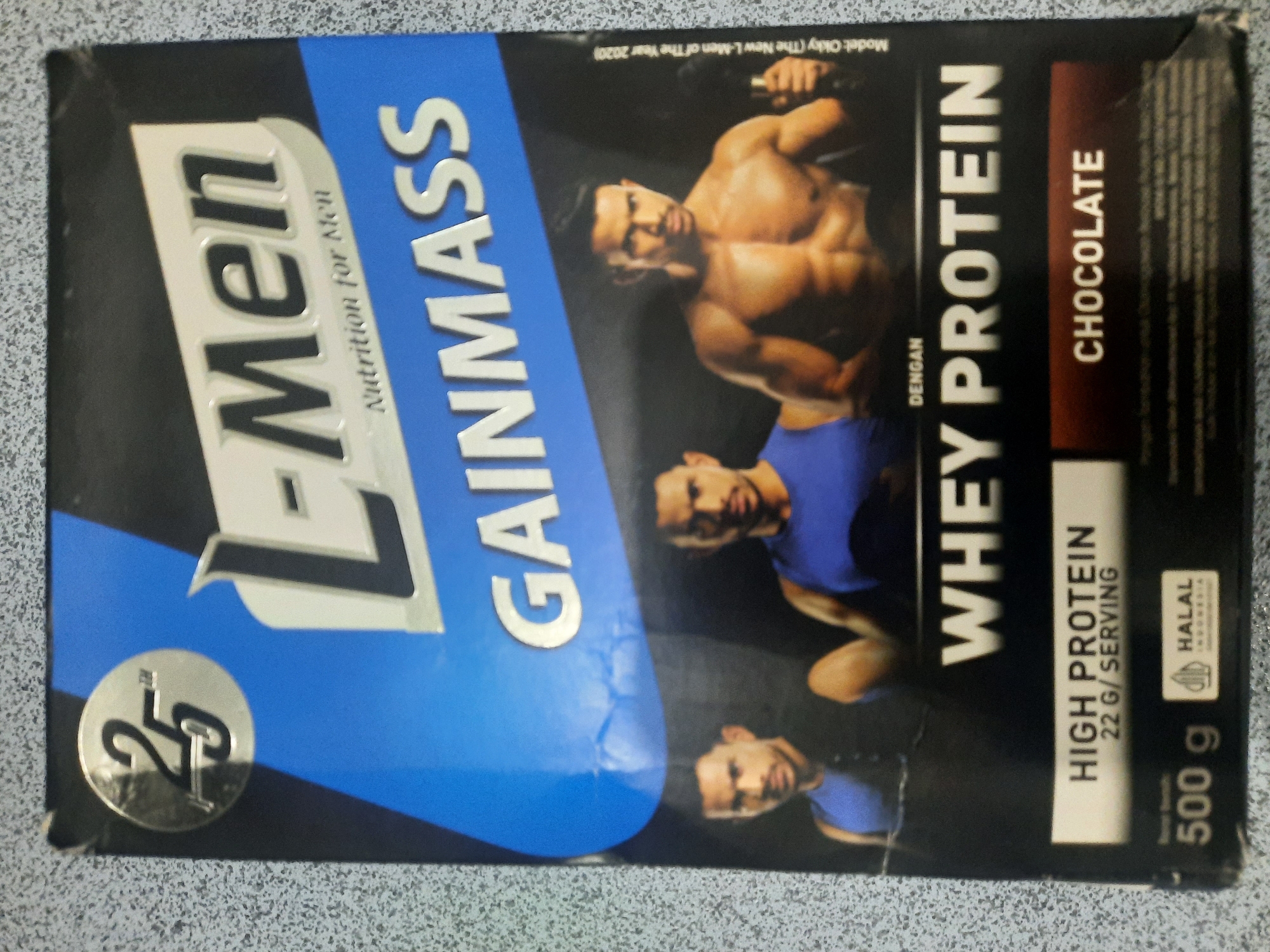 The image shows the front of a L-Men Gainmass chocolate flavored whey protein product. The product packaging features three muscular men, the L-Men logo, and text indicating "High Protein 22 G/Serving". The packaging also includes a "Halal Indonesia" certification and specifies a net weight of 500 grams. There is no information in the image or its crops that allows for the determination of its location in Lhokseumawe, Indonesia.
