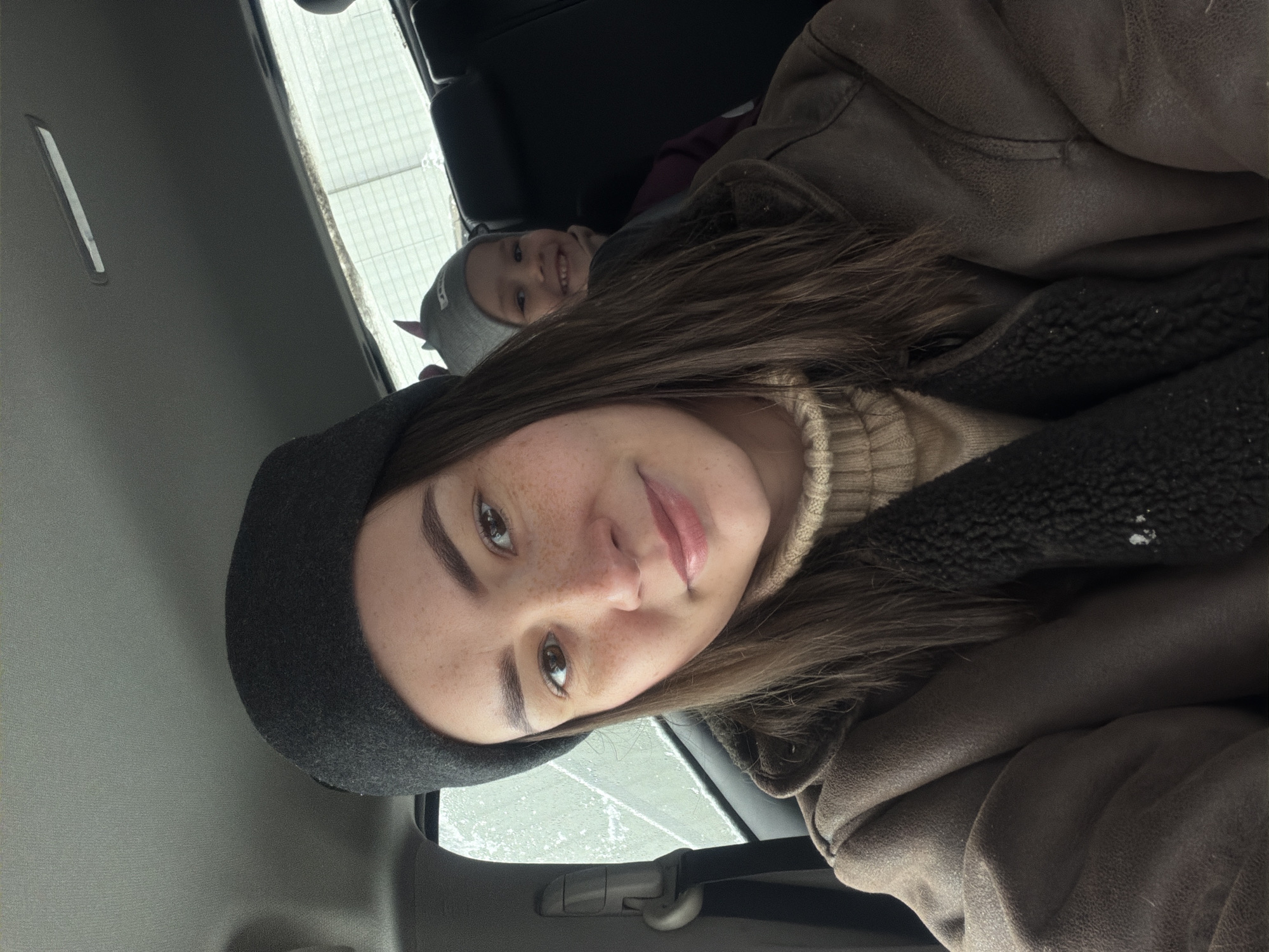 A selfie of a woman wearing a dark grey beret, brown jacket, and beige turtleneck sweater in the backseat of a car. A young child wearing a grey hat with horns is in the background, also in the backseat, smiling. The car window shows a view of snow. The location is likely Chaikovsky, Russia.