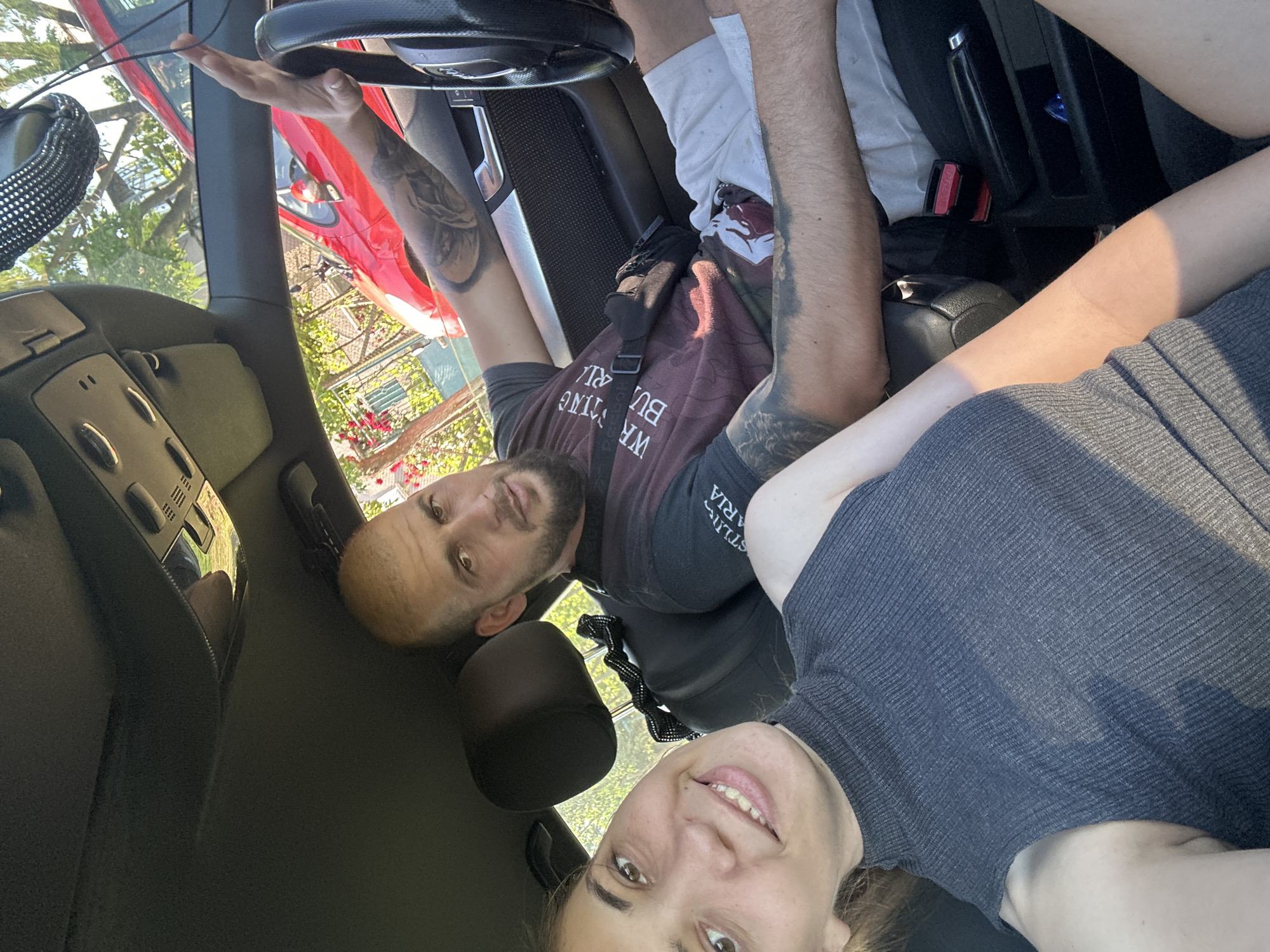 The image captures two individuals, a man and a woman, inside a car cabin, likely posing for a selfie. The perspective is from a low angle, looking upwards at the occupants. The original image is rotated 90 degrees clockwise, so the description assumes an upright orientation.
The man, situated towards the left (driver's side) of the frame, has a short beard and is looking directly at the camera with a calm expression. His arms are visibly tattooed, and he is wearing a dark t-shirt with some white text that partially reads "WE ARE BUILDING BUT..." or similar. A black strap from a bag or accessory crosses his chest. His right arm is extended upwards towards the car's windshield area.
To the man's right, the woman is smiling broadly at the camera. She has dark hair and is wearing a dark, ribbed sleeveless top.
The car's interior is dark-colored, showing parts of the dashboard, a steering wheel, and seat upholstery. Through the windshield, a bright, sunny outdoor scene is visible. A red car is parked directly outside, along with abundant green trees and foliage, and hints of buildings in the background. The bright natural light and visible shadows suggest it is daytime. The scene evokes a relaxed atmosphere, consistent with the specified location context of Rogozen, Bulgaria.