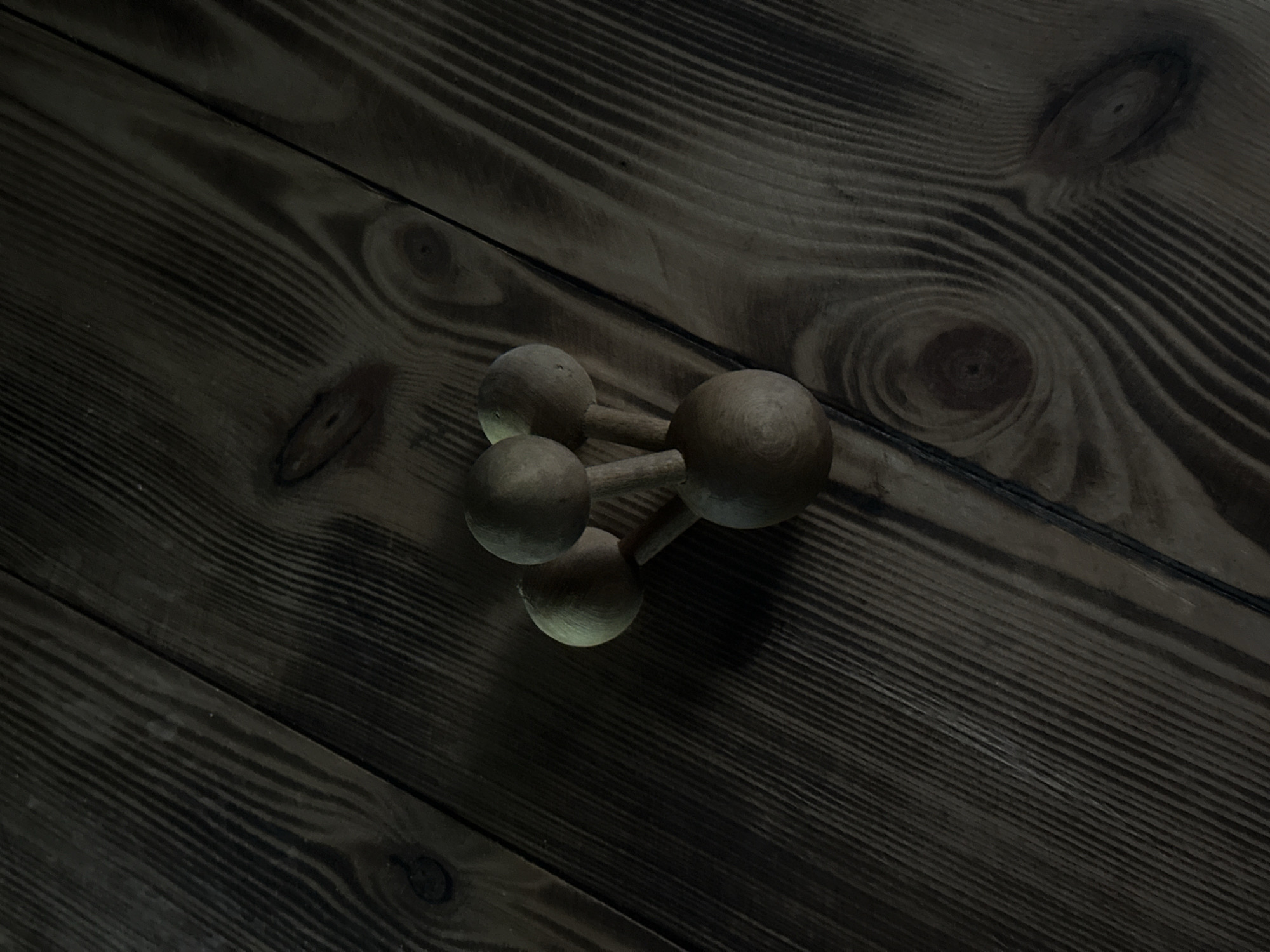An overhead, dimly lit, high-angle medium shot shows a wooden mushroom-shaped massage tool with four rounded knobs arranged in a cluster on a dark wooden surface. The wood has a prominent grain pattern and appears to be planks laid diagonally. The lighting is dim, casting soft shadows. There are no people or other objects visible in the frame. The location is not specified in the image.