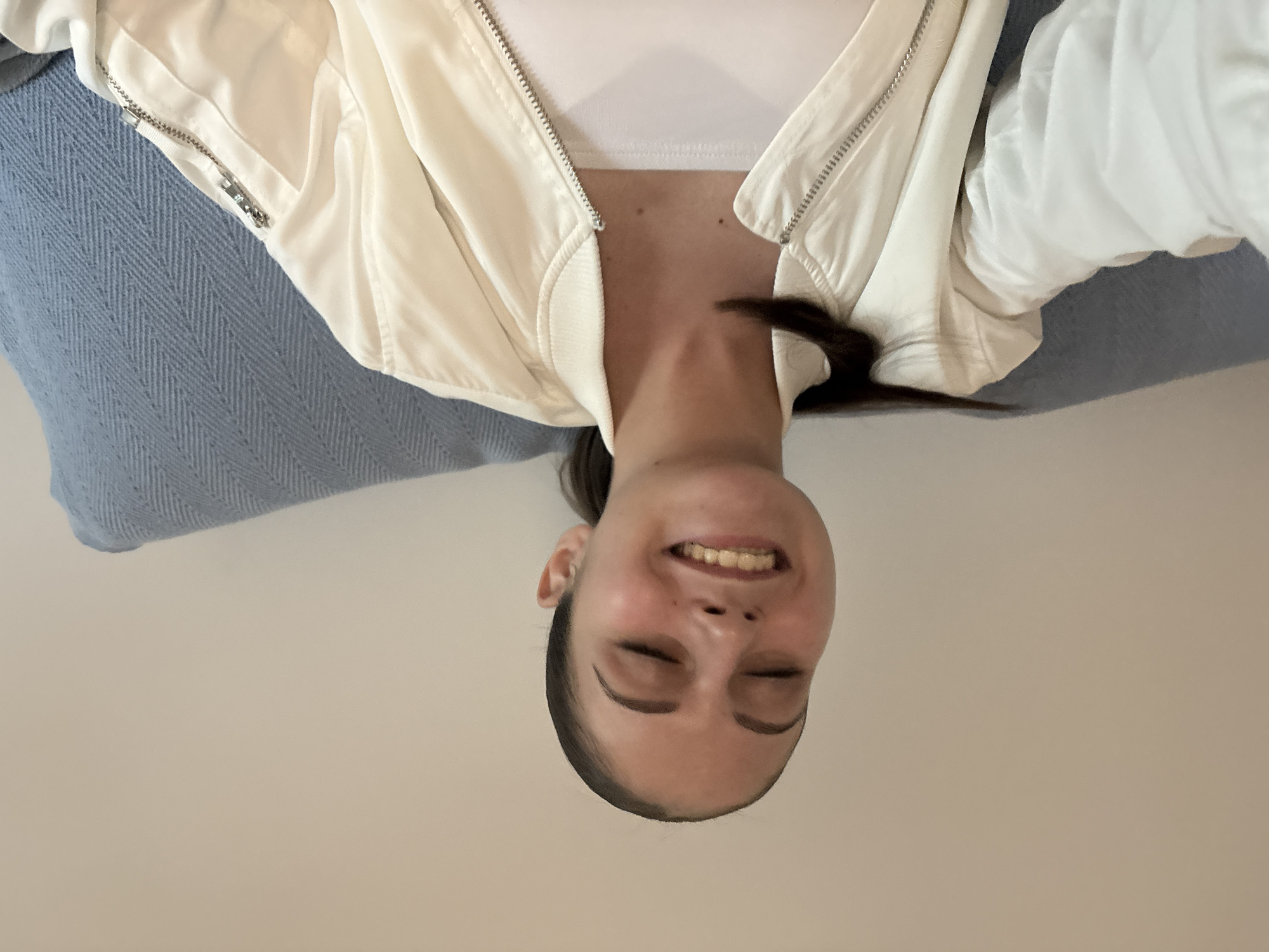 A person is pictured in a selfie-style image, with the camera positioned above and facing downward. The person has dark hair pulled back, closed eyes, and a smile revealing their teeth. They are wearing a white top and a partially zipped off-white jacket. A blue knitted fabric is visible to the left of the frame. The background is a plain, light-colored surface. The location and city are not discernible from the visual information provided.