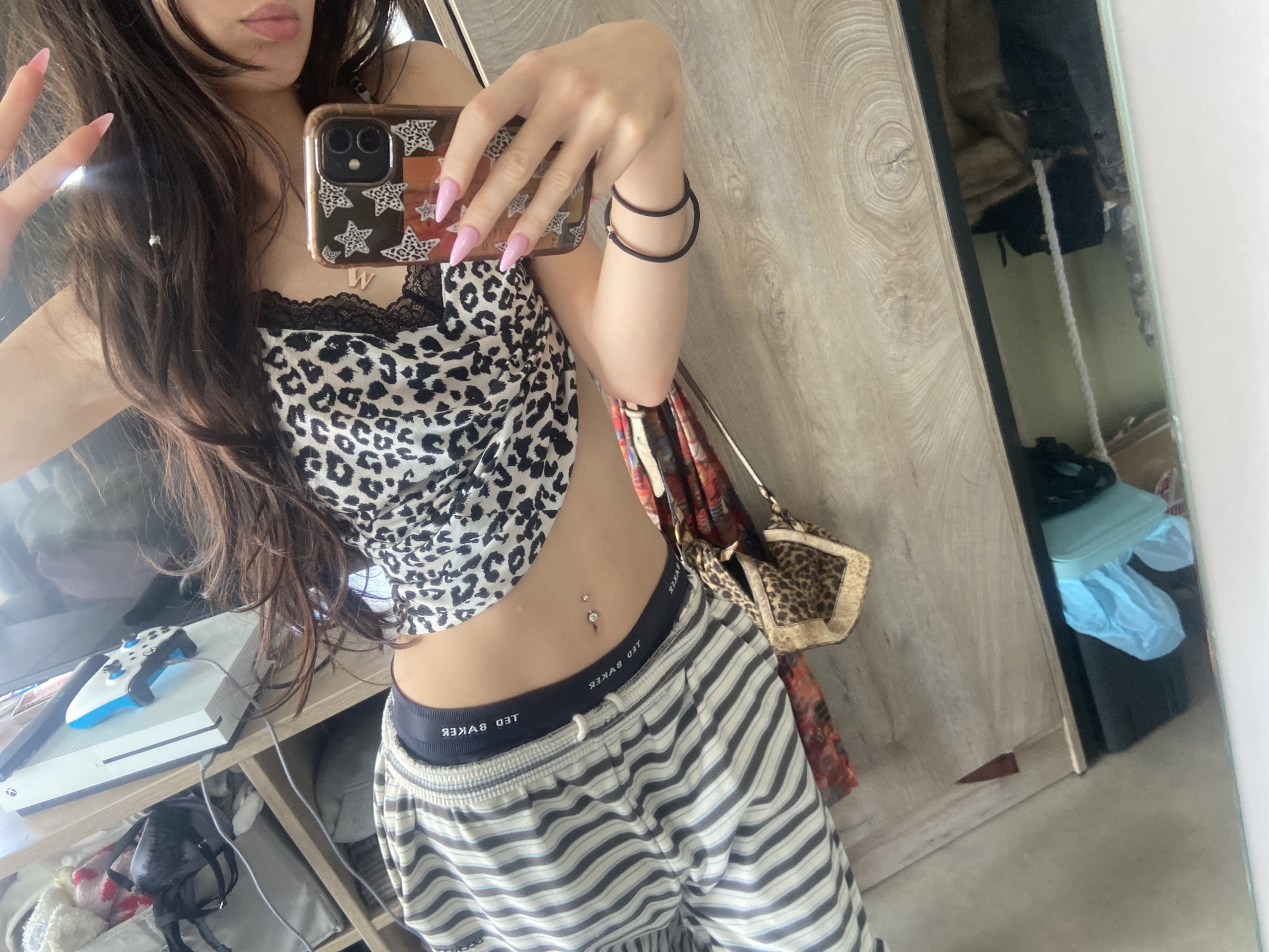 A person is taking a mirror selfie indoors. The person is wearing a leopard print tank top with black lace trim, striped pajama pants with a black waistband displaying "TED BAKER" in white text, and has two black elastic bands on their right wrist. The person's left hand is making a peace sign, while their right hand is holding a phone with a brown and black star-patterned case. A silver belly button ring is visible on the person's stomach. Behind the person, there is a television with an Xbox and controller on a shelf. To the right, there is a wardrobe with a purse hanging on the door. The environment is domestic, but there is no clear indication that this is in the city of London, United Kingdom.