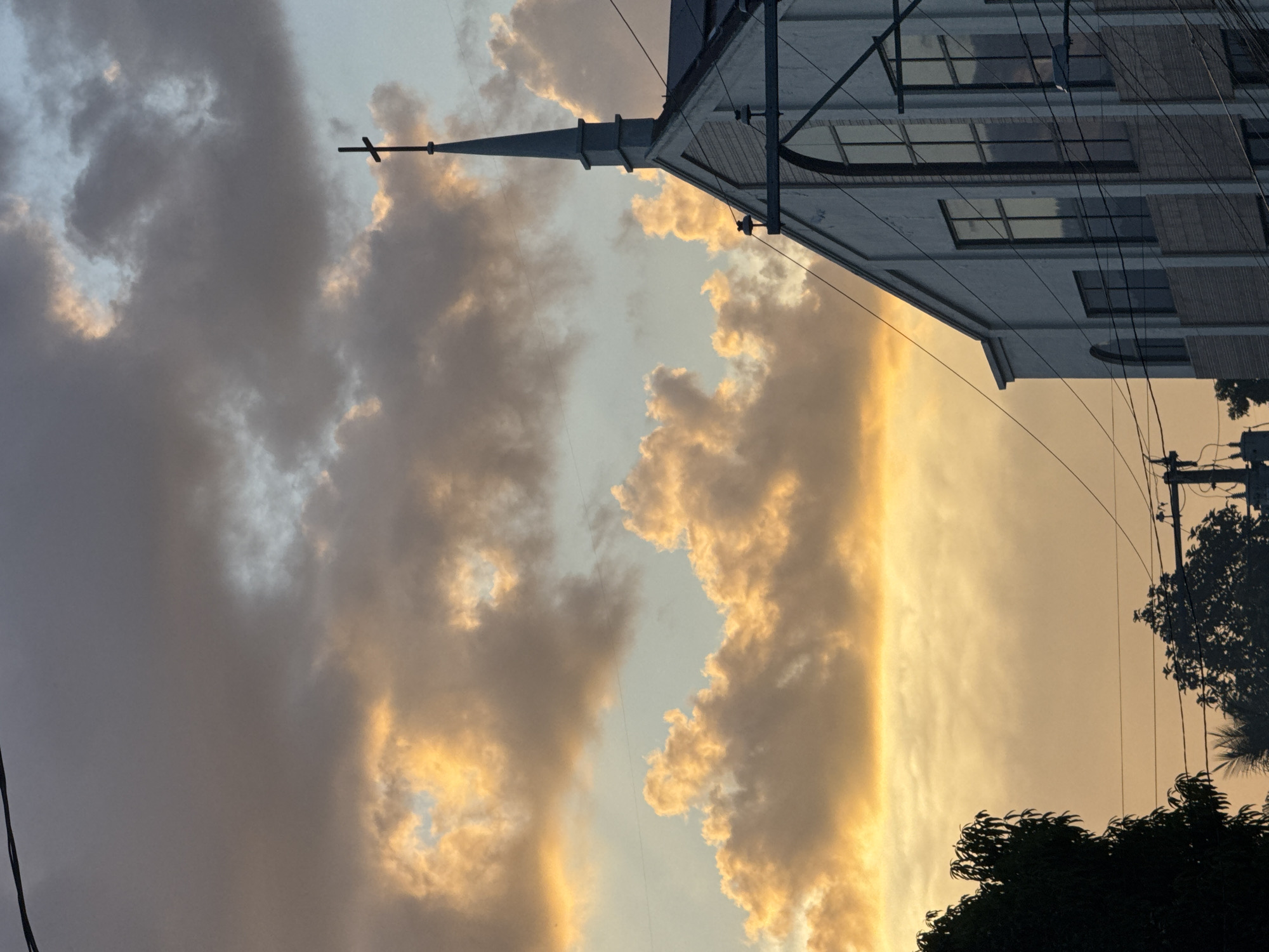 The image captures a view of a church steeple against a backdrop of dynamic, sunset-lit clouds. The church structure is partially visible on the right, appearing as a light-colored building with visible windows. Power lines are strung across the upper-right portion of the frame. The sky is filled with clouds ranging from dark gray to luminous yellow, suggesting either the beginning or end of the day. Some trees are visible in the lower right. This is likely in Davao City, Philippines.