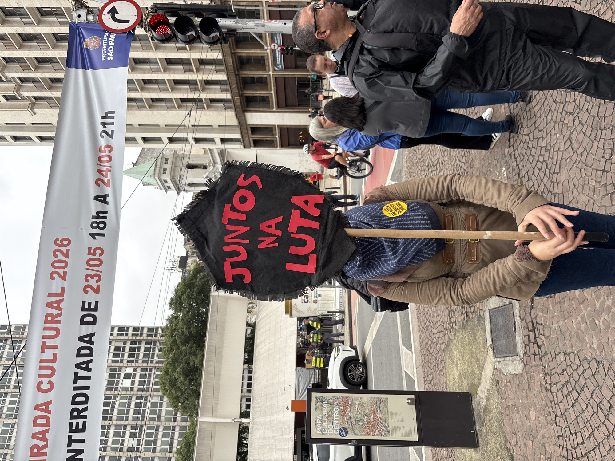 The scene depicts an urban street in São Paulo, Brazil, with several key elements. A banner promoting "RADA CULTURAL 2026" is prominently displayed. A woman holds a black, fringed sign with the phrase "JUNTOS NA LUTA" (Together in the Fight) written in red. Her lower torso and arms are visible; she wears a brown jacket. In the background, other people are present, including a man in a black jacket and glasses, and a cyclist. Traffic lights are visible above. Police officers stand guard near a white car, suggesting a controlled environment or event. A map titled "MAPA CULTURAL DO CENTRO" is positioned near the woman. The street is paved with a textured, cobblestone-like pattern. The text on the banner indicates dates "23/05 18h A 24/05 21h" and also includes "PREFEITURA SÃO PAU," signifying the city government's involvement. The street is closed to traffic, as signified by the "interditada" in the banner, the red traffic lights, and the position of police.