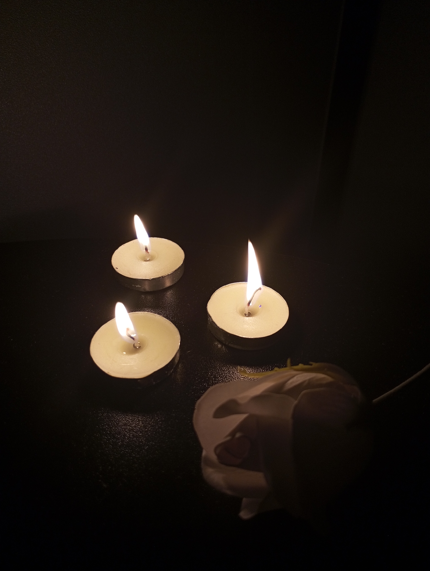 The image shows three lit tea light candles and a white rose on a dark, possibly black, surface. The background is dark and indistinct, with a vertical dark line possibly indicating an edge or corner. The overall lighting is dim, emphasizing the flames of the candles. There are no people visible in the image. The photo does not give any indication about the location being in the city of Pervomaysky, Russia.