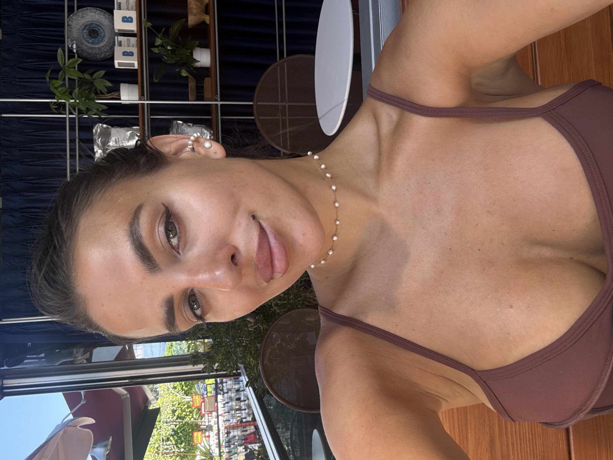 A woman with dark hair pulled back and dark makeup is taking a selfie outdoors. She is wearing a brown bikini top and a pearl choker necklace. She has a pearl earring in her left ear. The background shows a blue curtain, shelves with plants and boxes, and some outdoor seating with a table. The image appears to be taken at a cafe or restaurant in Fethiye, Turkey, given the presence of outdoor seating and what appear to be price tags visible in the blurred background to the left.
