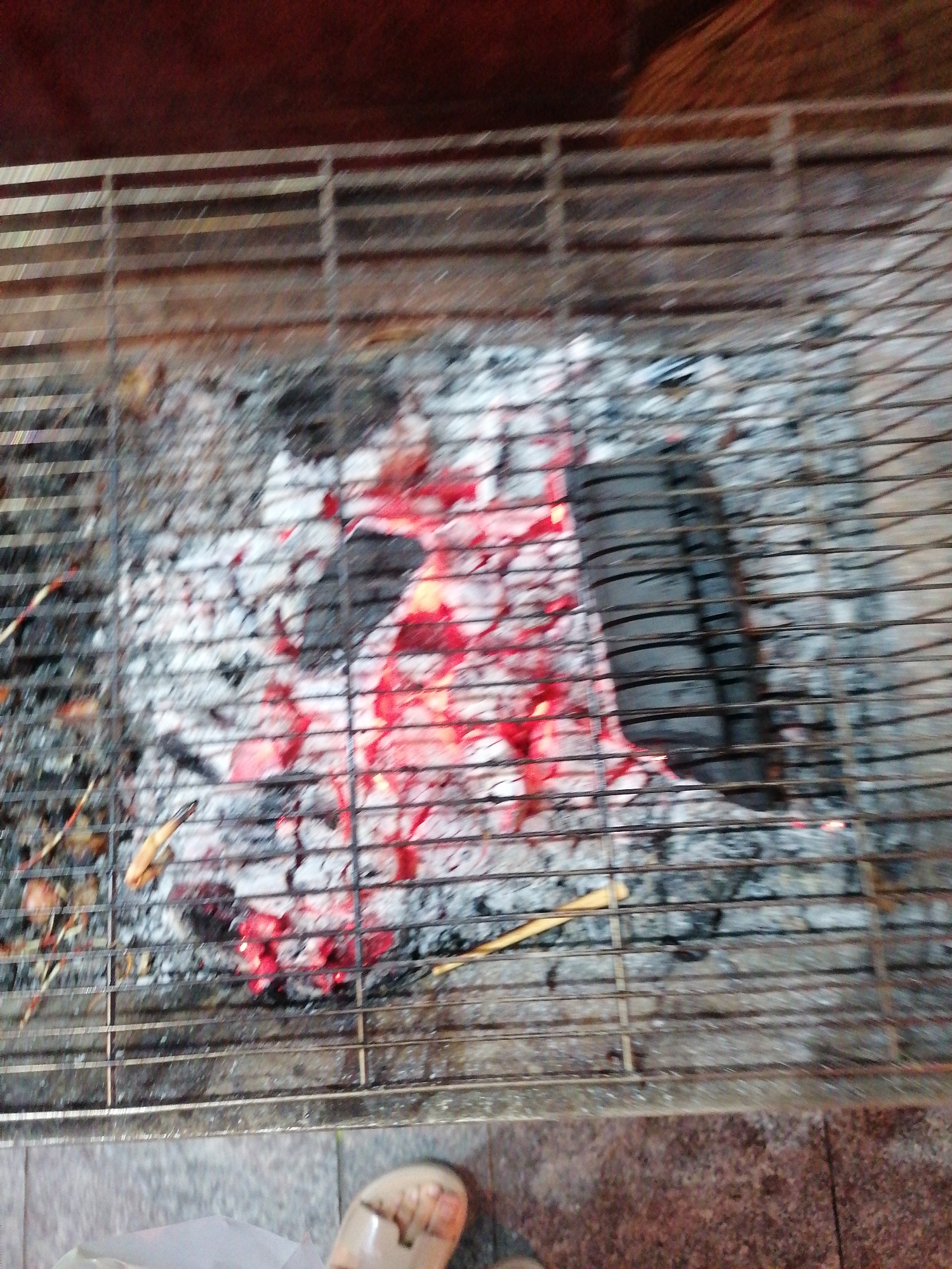 A top-down shot shows a metal grill with hot coals underneath. The coals are glowing red in the center, surrounded by gray ash. A dark, partially burned briquette is visible on the right. Below the grill, a person's foot wearing a sandal can be seen. The environment appears to be a patio or outdoor area. It's not possible to determine the precise location within Nha Trang Ward, Vietnam, based on the image.