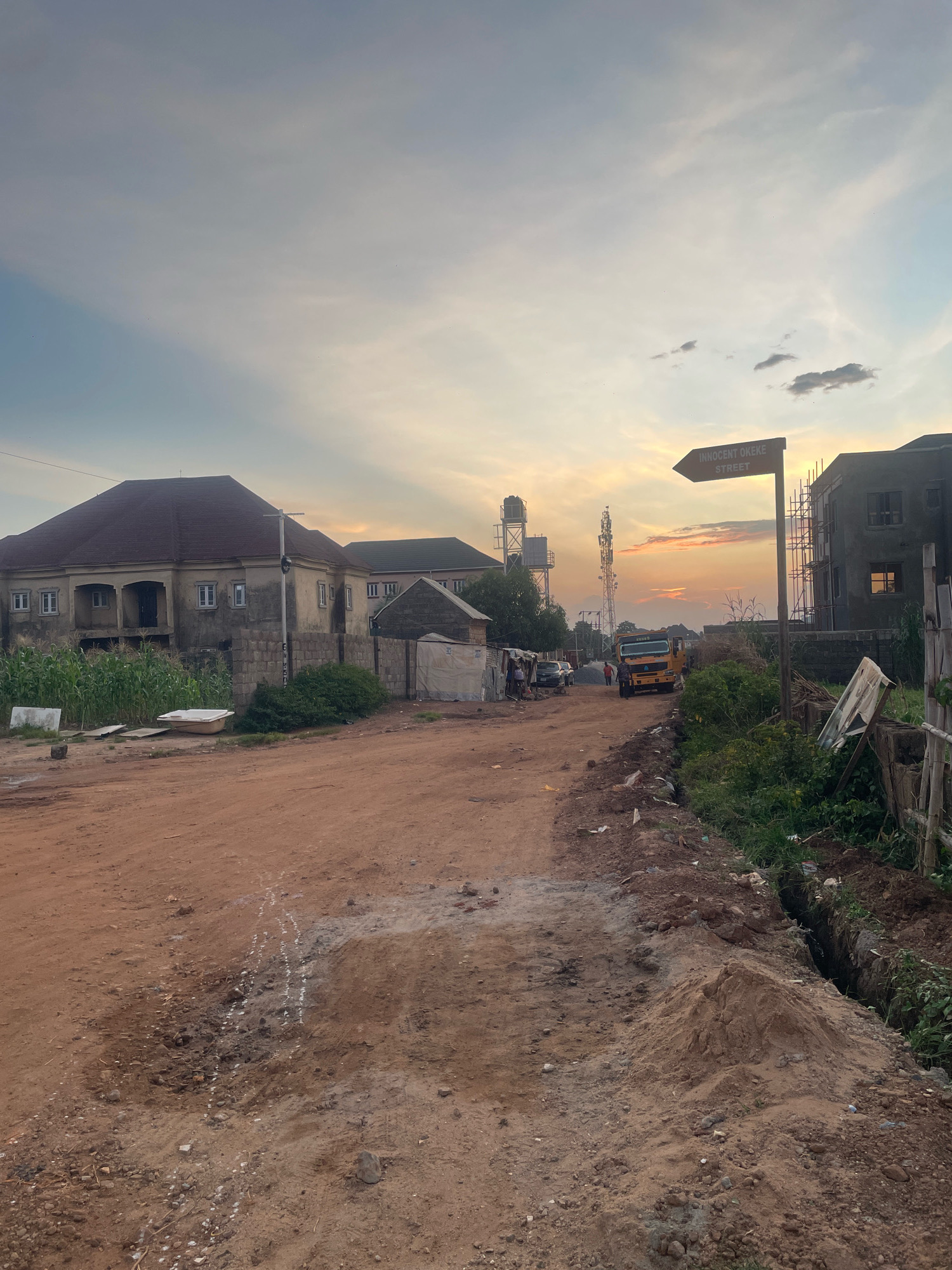 The image depicts a street scene in Abuja, Nigeria, characterized by unpaved roads and ongoing construction. In the foreground, a dirt road is visible, with piles of dirt and some white markings on its surface, suggesting recent activity or preparation for construction. To the left, a large, multi-story building with a brown roof and several windows is partially obscured by corn stalks. Adjacent to this is a smaller, more rudimentary structure covered with a tarp.
Further down the road, a yellow truck is parked, and several people are standing nearby. Other buildings, some appearing to be under construction with scaffolding, are present in the background. A prominent signpost reads "INNOCENT OKEKE STREET" with an arrow pointing to the right. The sky is overcast with a warm glow, indicating either sunrise or sunset, with some clouds visible. Water tanks and a communication tower are also discernible in the distance. The overall environment appears to be a developing urban or peri-urban area.
