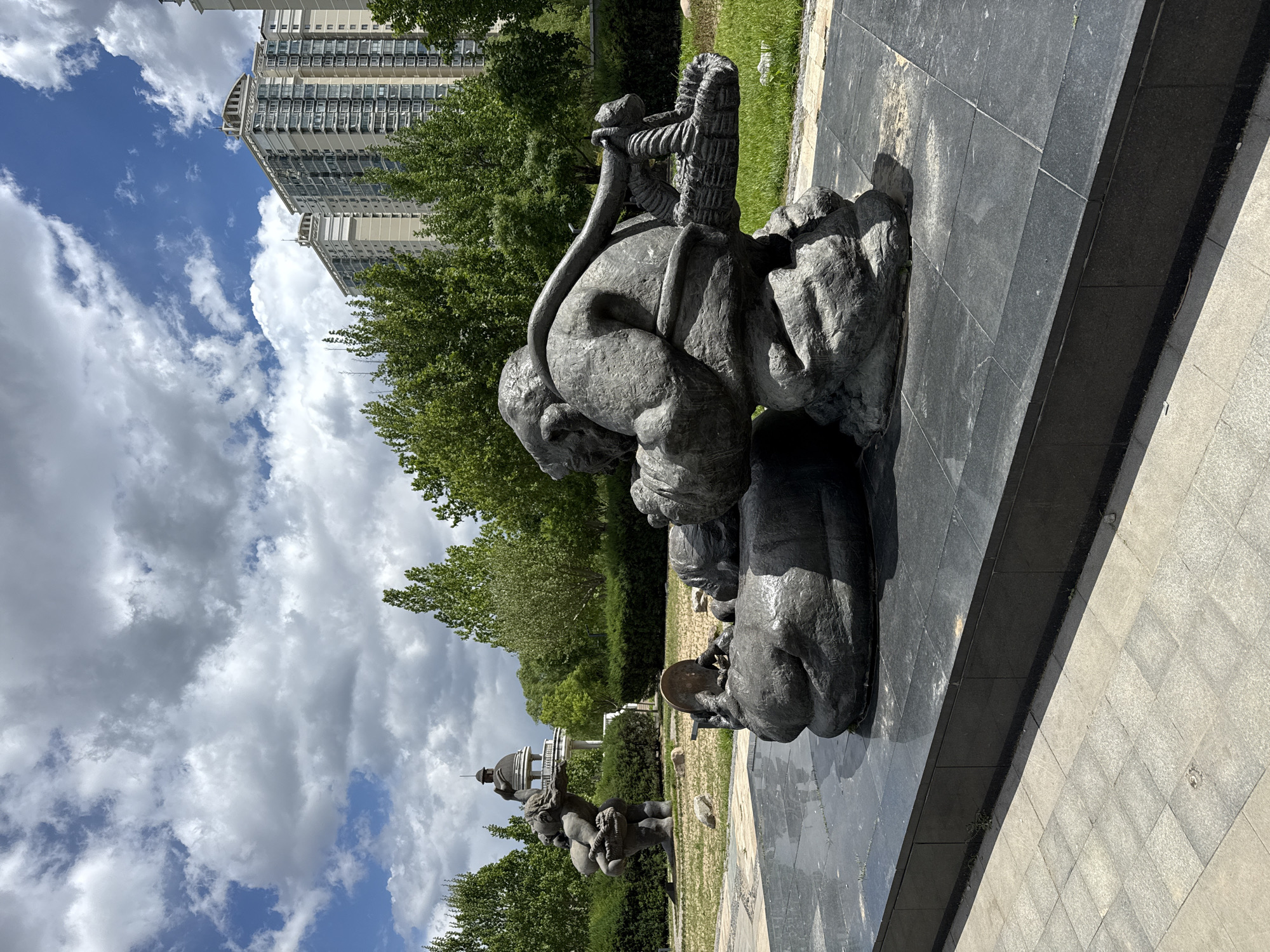 The image shows a bronze sculpture of people on a paved area. The sculpture depicts several figures, including someone carrying a load on their back and another figure holding a circular object. In the background, there are trees, a tall building, and a gazebo-like structure. The sky is partly cloudy. The location is Changping District, China.