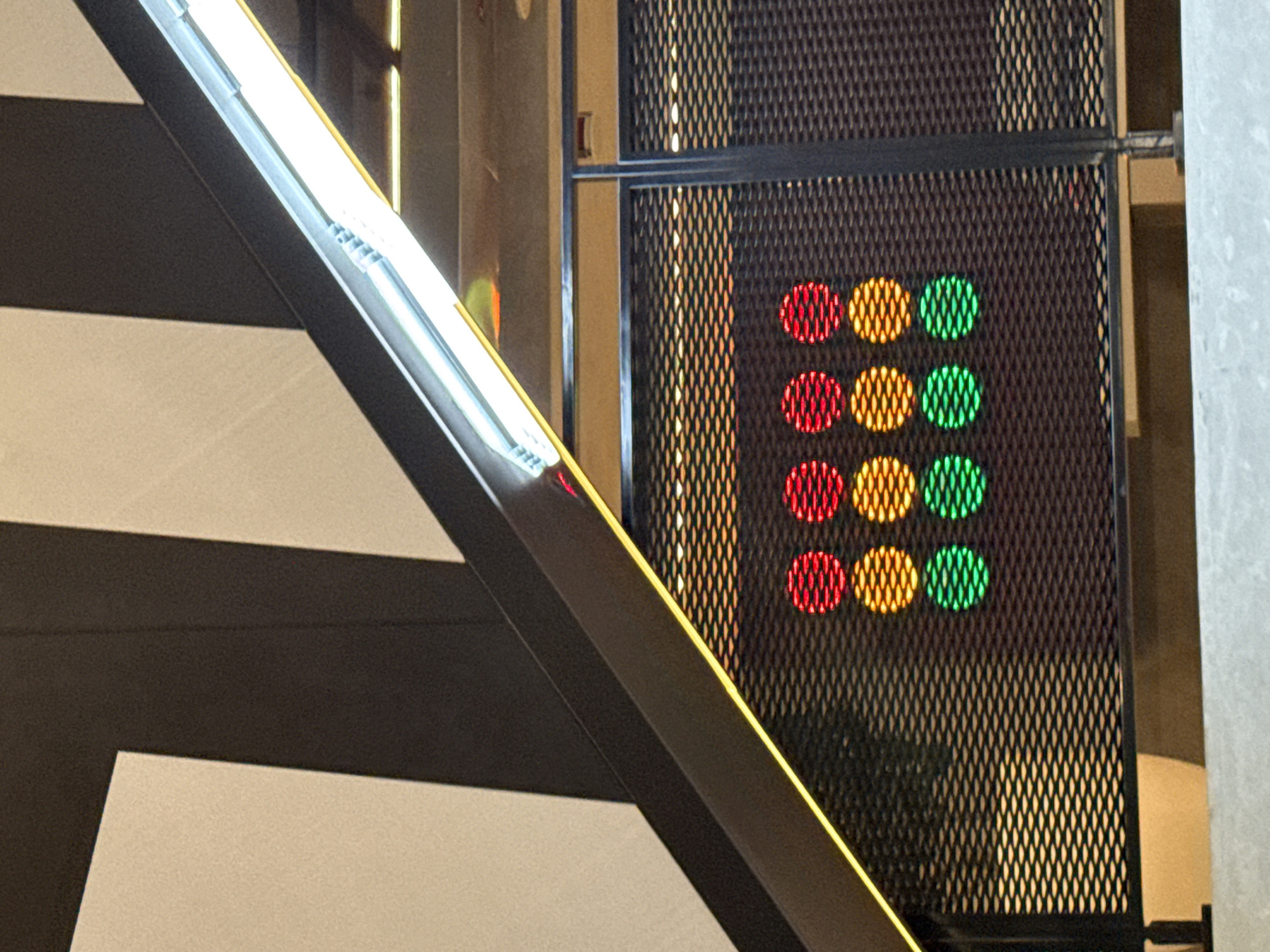 An eye-level, medium shot captures a close-up view of a metal grate illuminated by a linear fluorescent light fixture. Behind the grate, a series of circular lights are visible in a grid pattern. The lights are arranged in three rows of four, with each row displaying a different color: red at the top, orange in the middle, and green at the bottom. The overall scene appears to be indoors, possibly in a commercial or public space, with a large, dark geometric shape dominating the upper left portion of the frame. The image does not provide any specific indicators of its location within Dubai, United Arab Emirates.