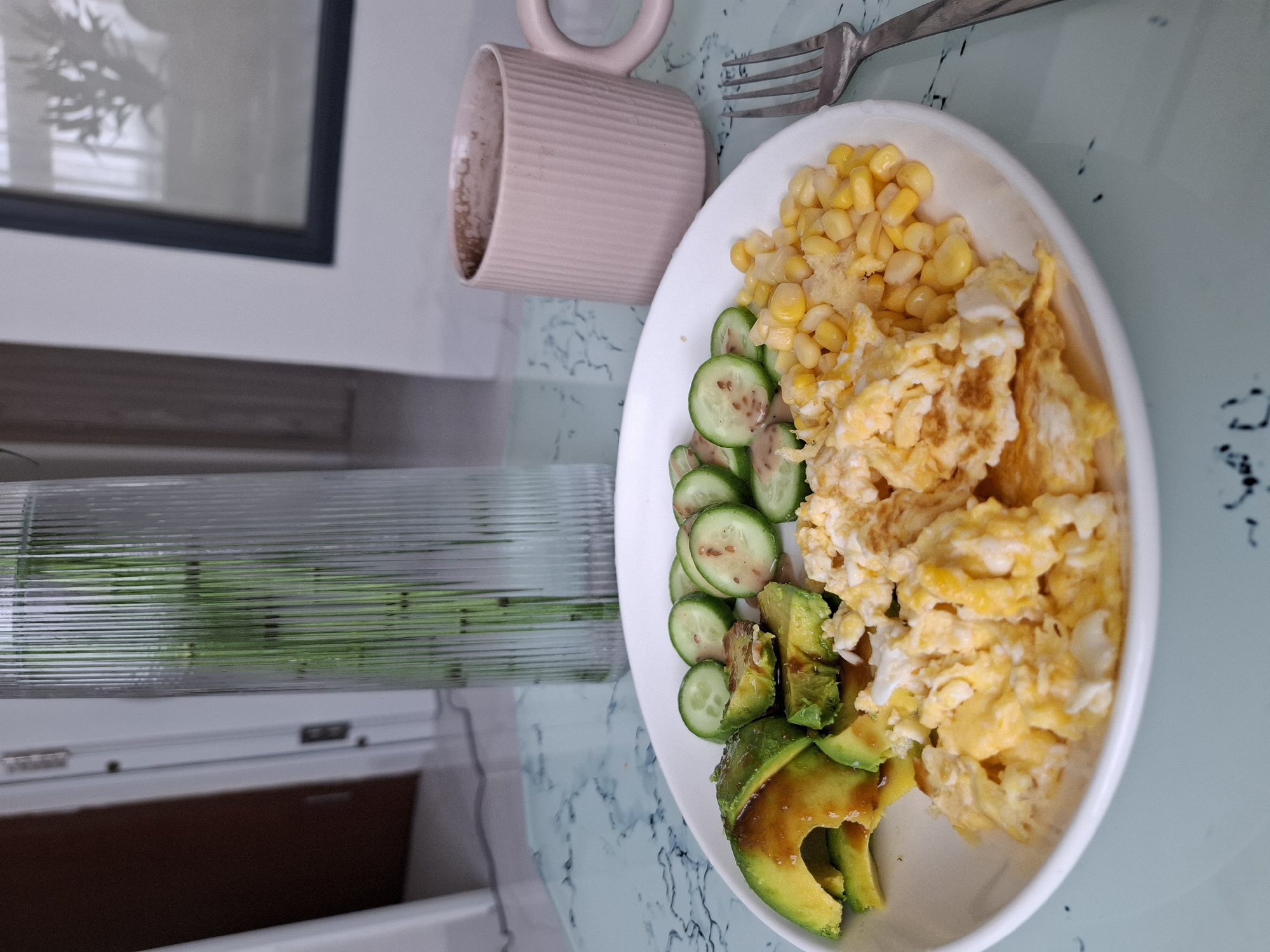 This is a close-up, eye-level, outdoor, natural light photograph of a breakfast spread on a marble-patterned table. The focal point is a white plate containing scrambled eggs, corn, sliced avocado drizzled with sauce, and sliced cucumbers. To the right of the plate is a silver fork. In the background, to the right, is a pink ridged mug filled with a beverage. Behind the mug and to the left is a tall, clear glass vase with green stems. The table surface has a light blue and white marble pattern. The background appears to be an indoor setting with a blurred doorway and a framed picture on the wall. The scene suggests a meal being served or eaten, with no people visible. The location is identified as Nha Trang Ward, Vietnam.