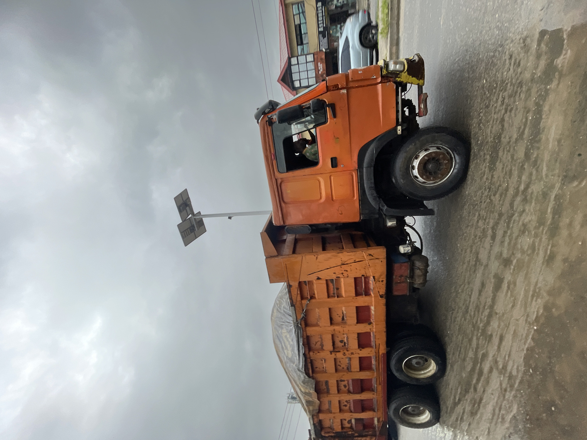 The image shows an orange dump truck parked on a wet asphalt street in Ado, Nigeria. The sky is overcast. A utility pole with street lights is visible behind the truck's cab. A silver car and buildings are visible in the background. The truck bed is partially covered with a grey tarp. A person is visible inside the cab.