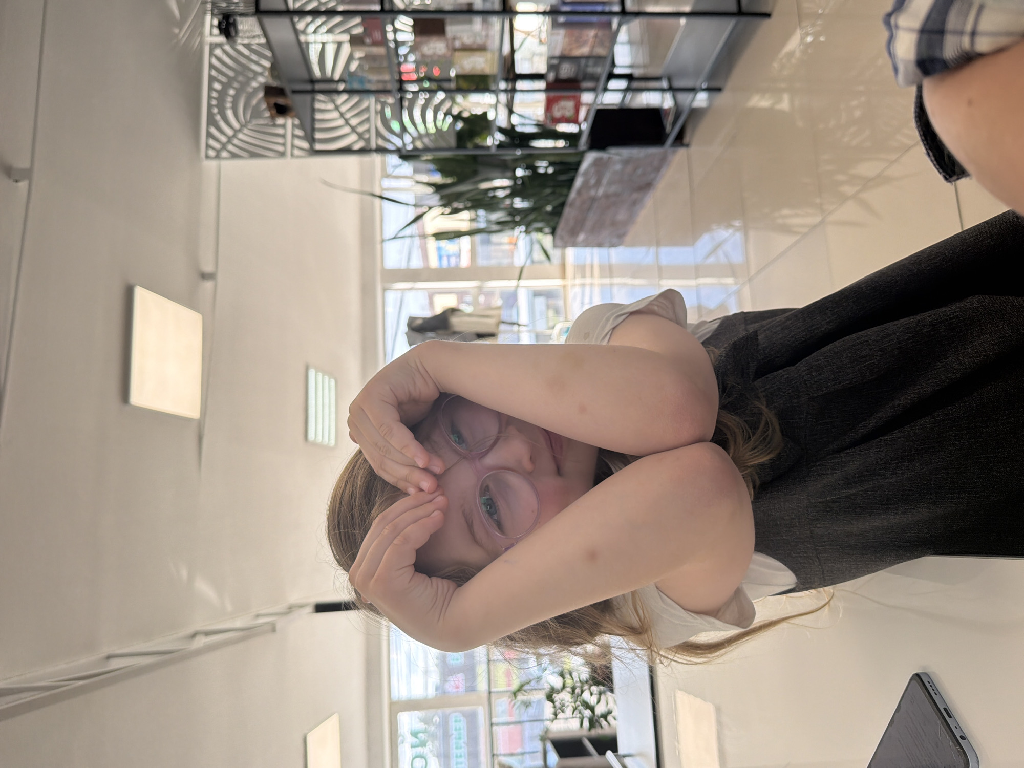 The image, when correctly oriented, shows a young person, likely a child or pre-teen, with light brown hair and pink-rimmed glasses, looking directly at the camera. Their hands are playfully positioned over their face, forming a heart shape and partially obscuring their eyes, giving a shy or whimsical expression. They are dressed in a light-colored top and a dark skirt or trousers. Part of another individual, possibly the photographer, is visible on the right, wearing a blue and white checkered shirt.
The scene is an interior space, characterized by bright white walls and a reflective, light-colored floor, suggesting a modern and clean environment like a cafe, shop, or public library. In the background, a distinctive multi-tiered metal shelving unit with an intricate, web-like design holds various items, including small potted plants and what appear to be books or merchandise. Large windows behind the shelving unit offer a glimpse of an urban exterior with buildings and bright daylight.
Notable details include a smartphone with a light blue case and a cracked screen resting on a white surface in the foreground on the right. Ambient lighting is provided by square and rectangular fixtures on the walls and ceiling. Visible through the window, faint Cyrillic text, possibly "ТЕЙЛЕР", is discernible on an exterior sign or reflection, consistent with the specified location of Chaikovsky, Russia. The image captures a candid, daytime moment.