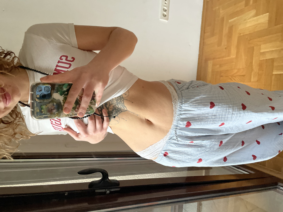 The image depicts an indoor scene in Boreti, Montenegro. A partially visible individual, presumed female, with light curly hair and a faint smile, is centered in the frame. The subject is wearing a white short-sleeved crop top displaying red, inverted text on the chest area, and light blue and white striped pajama pants with red heart patterns. White lace-trimmed underwear is visible above the pajama pants. A dark, beaded necklace is worn, and an intricate tattoo is visible on the lower left abdomen. The individual holds a mobile phone with a clear, patterned case in both hands, positioned as if taking a mirror selfie. The immediate environment includes a dark brown door or window frame with a black handle on the left, a plain white wall with a double electrical outlet on the right, and light-colored wooden parquet flooring laid in a herringbone pattern.