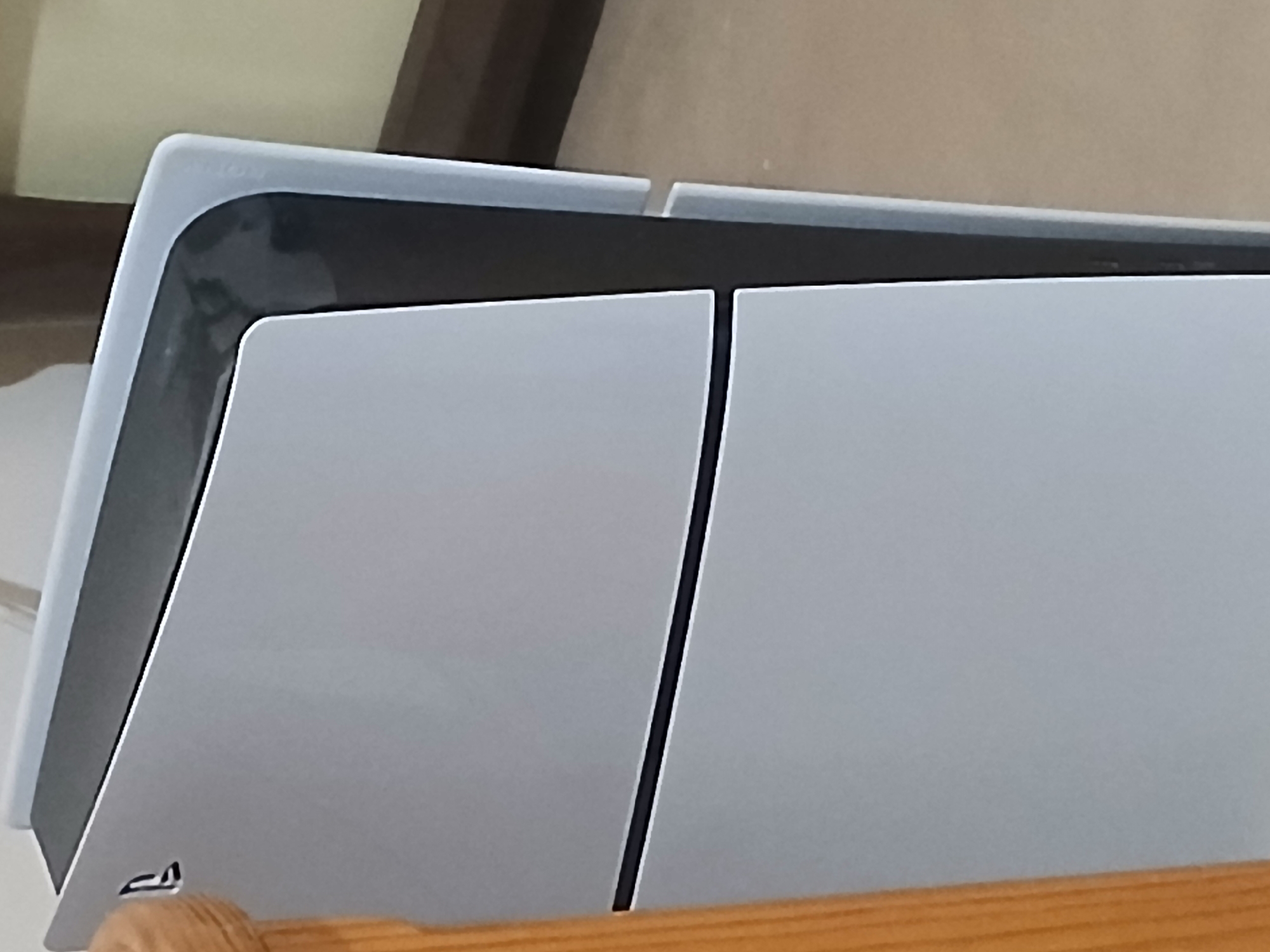 An extreme close-up shot shows the side of a white PlayStation 5 console standing vertically, positioned next to a wooden railing. The console has a sleek, curved design with a matte white exterior and a black inner section. A subtle black line divides the white panels of the console. The background is blurred, with hints of a light-colored wall and floor, and a dark wooden surface. The image does not provide any information about the city of Dnipro, Ukraine.