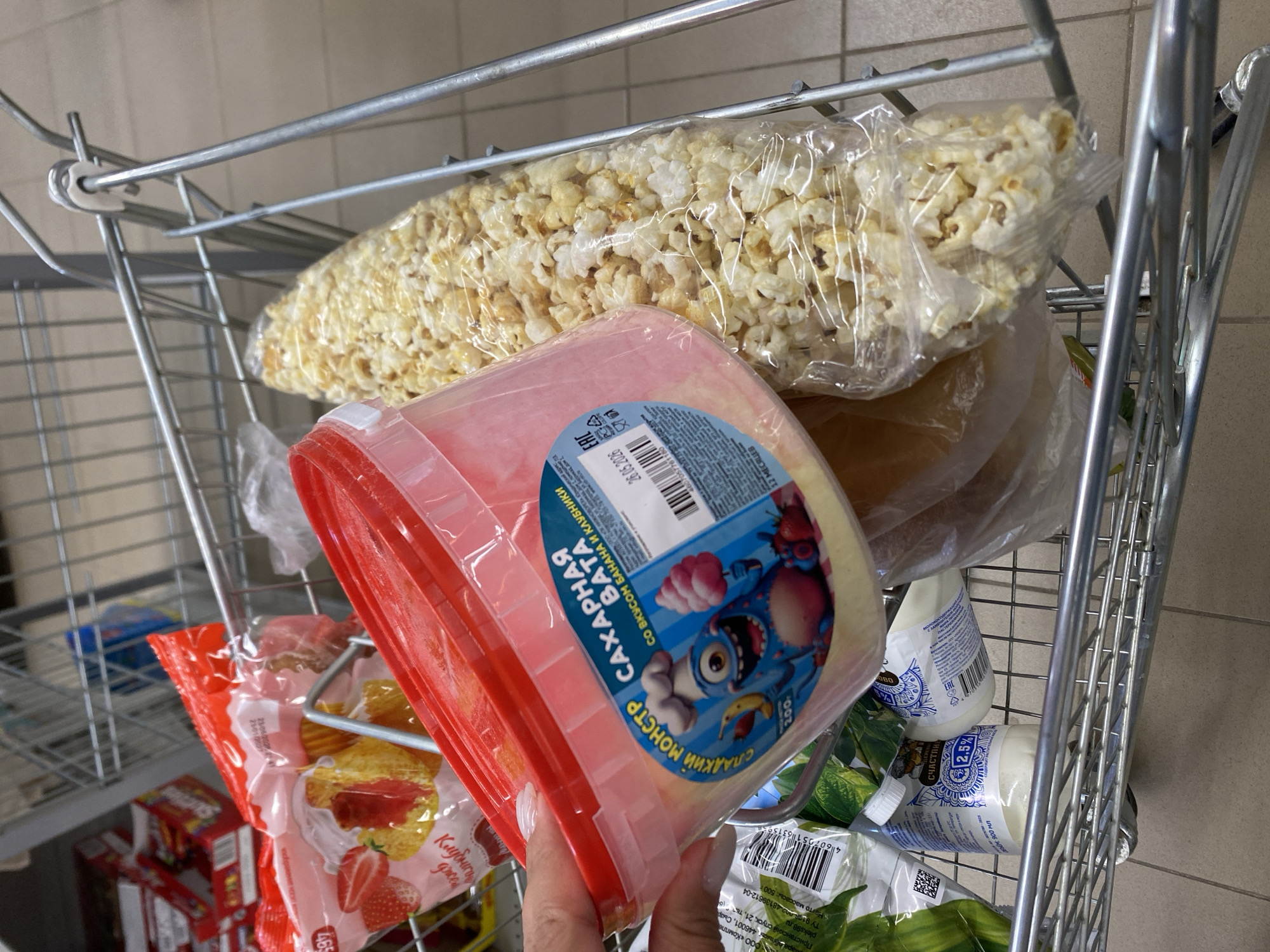 The image shows a shopping cart filled with various grocery items. In the foreground, a person's hand with manicured nails is holding a pink plastic container of cotton candy. The container has a label with blue text and a cartoon monster character. Next to the cotton candy is a large, clear plastic bag filled with popcorn. In the background, other items are visible, including a red bag with strawberries and cake, and several white bottles with blue and white labels. The shopping cart itself is made of metal and is positioned on a tiled floor. The overall scene suggests a person is shopping for snacks. There is no information available about the specific location in Kushnarenkovo, Russia.