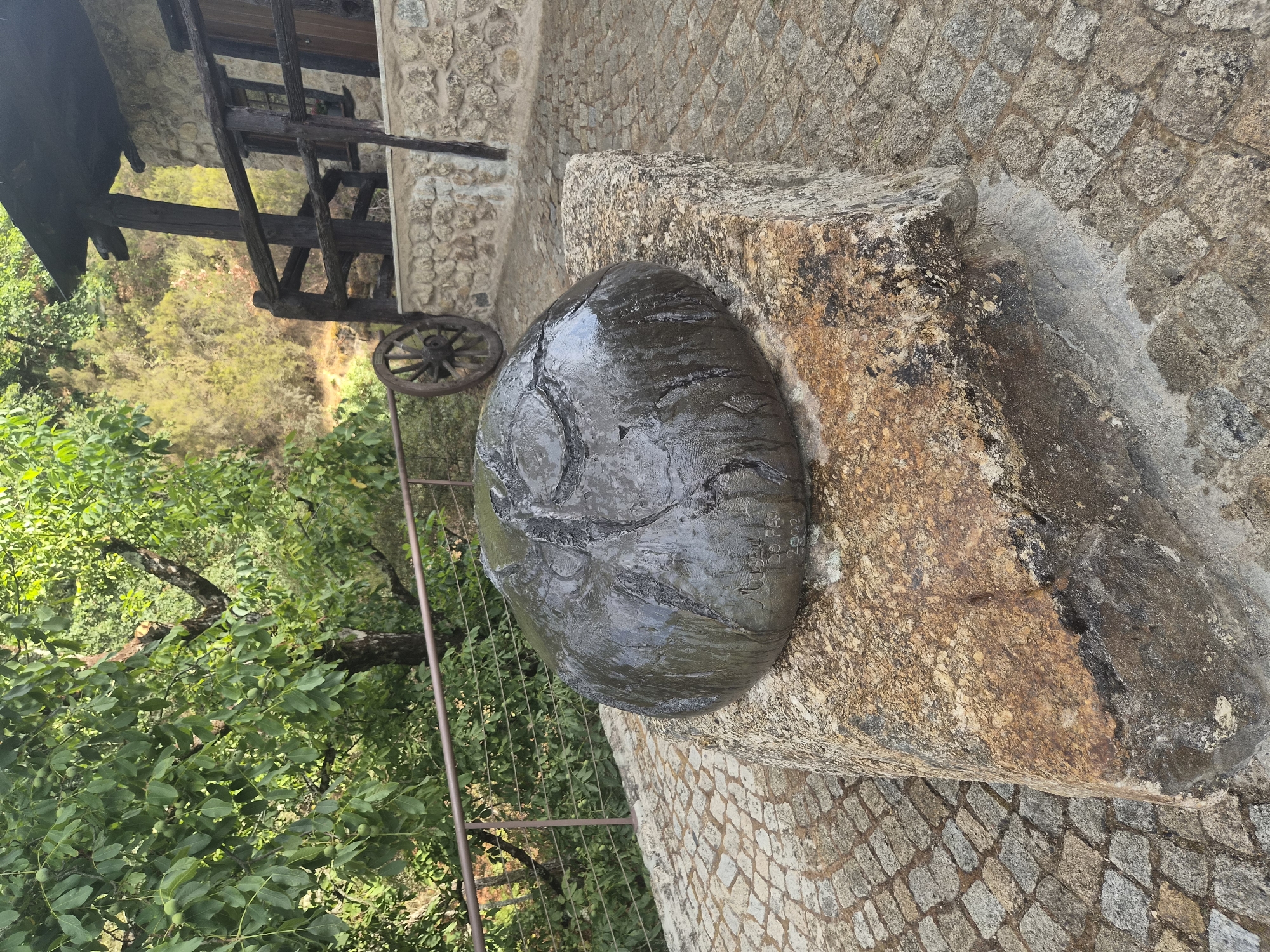 The image shows a large, dark, rounded sculpture placed on top of a rough, rectangular stone. The sculpture has a textured surface and appears to be wet, with reflections visible. There is some text inscribed on the sculpture, including what looks like "A Moreau", "Do Prado", and "2002". The sculpture and its base are situated on a cobblestone pathway. In the background, there is a wooden railing and a stone building with a thatched roof, suggesting a rustic or historical setting. Lush green trees and foliage are visible, indicating an outdoor environment. A vintage wooden cartwheel is mounted on the stone wall of the building. The overall scene suggests a park or a courtyard in a rural or semi-rural location. This sculpture is located in the Municipal Garden of Seia, Portugal, specifically in the area known as the "Jardim da Assunção".