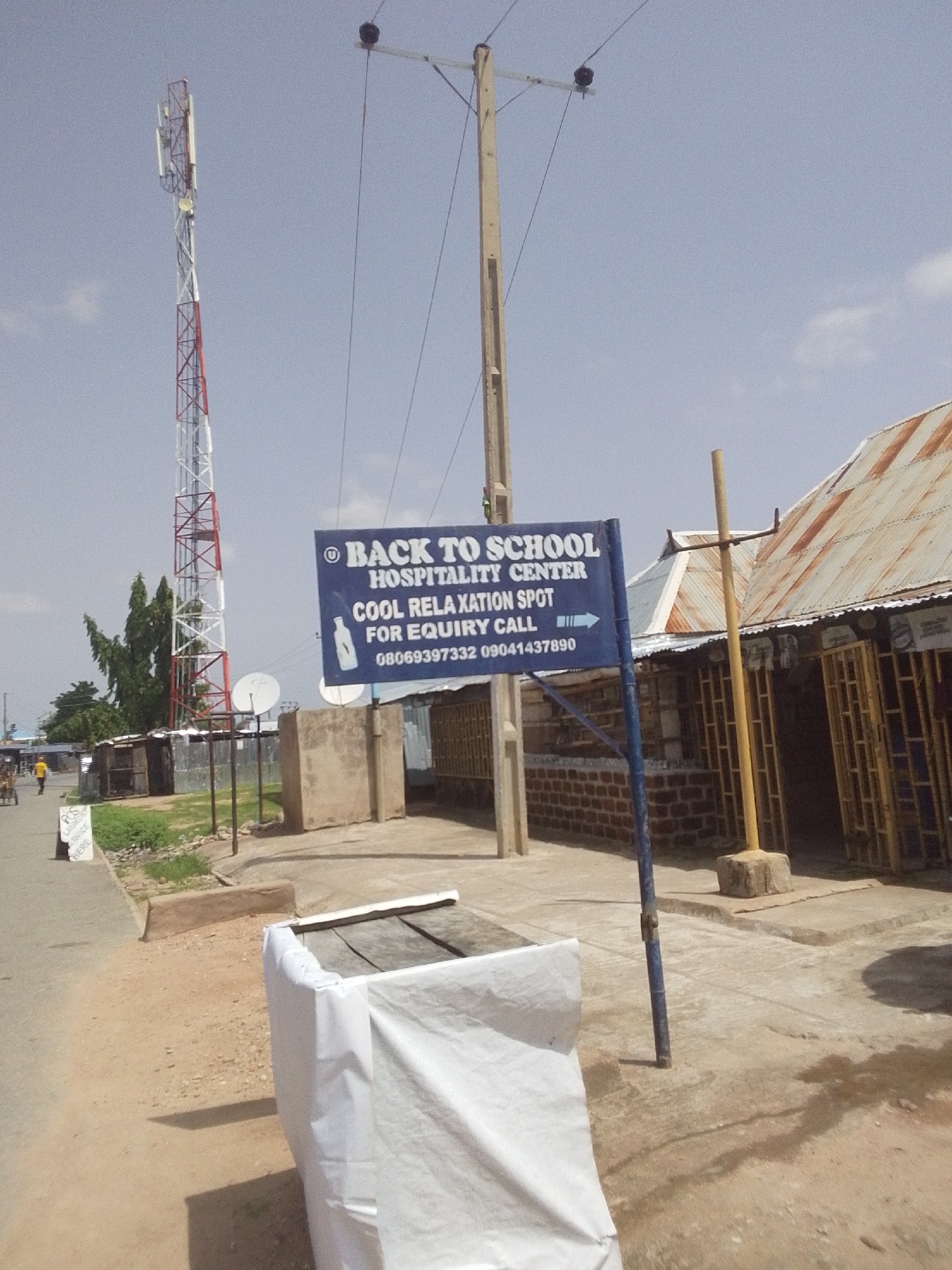 A street-level outdoor medium shot captures a sign that reads "BACK TO SCHOOL HOSPITALITY CENTER COOL RELAXATION SPOT FOR EQUIRY CALL 08069397332 09041437890." The sign is blue with white lettering and is mounted on a blue pole. Behind the sign, a red and white telecommunications tower stands tall against a pale blue sky with some clouds. To the right of the sign, a building with a corrugated metal roof and brick walls is visible. In the foreground, a white tarp covers an object, possibly a bench, on a dusty ground. A few small green plants grow near a concrete curb. In the distance, a person walks along the street, and a cart is being pulled by another person. The scene appears to be in a developed area with electrical poles and wires. The lighting suggests it is daytime, and the weather is clear. The overall impression is of a local business in a community setting in Jalingo, Nigeria.