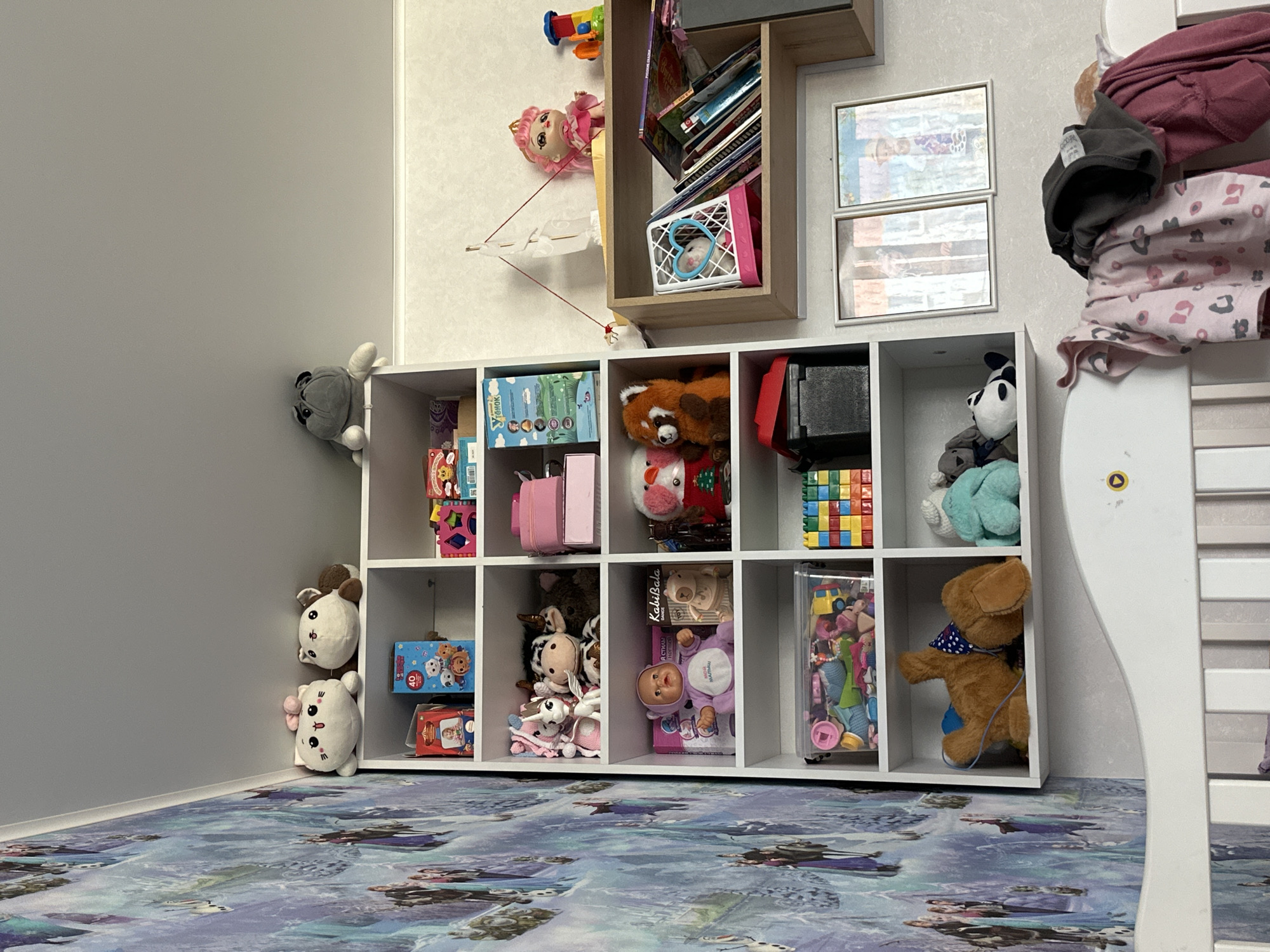 The image displays a white bookshelf filled with children's toys and books, positioned against a wall adorned with a Frozen-themed wallpaper. Two stuffed animal characters are on the top shelf, alongside what appears to be a plush pug. Below them, various toys and boxes are neatly arranged on the subsequent shelves. On the right side of the image, a smaller wooden shelf holds books, a doll with pink hair, and a toy train. Two framed pictures hang on the wall above this shelf. In the foreground, a white crib with bedding and clothing draped over it occupies the lower portion of the frame. The setting appears to be a child's bedroom.
