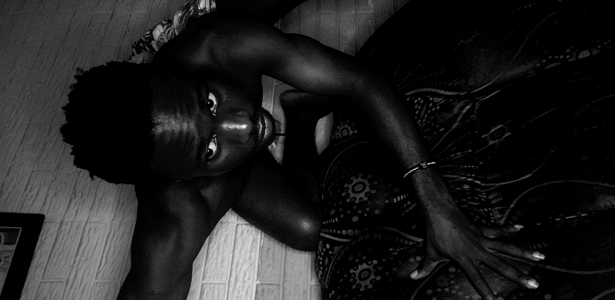 A low-angle, black and white photograph shows a dark-skinned person lying on a floor with patterned fabric. The person's head is tilted upwards, facing the camera. A hand rests on the fabric. To the left, a tiled floor is visible and a picture frame can be seen in the corner. It may be a photo taken at Narayi, Nigeria.
